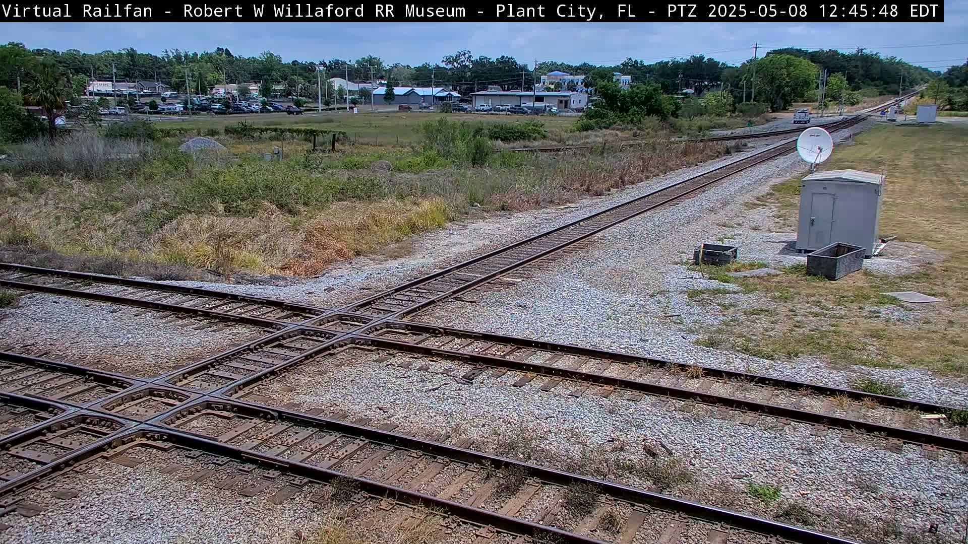 A railway crossing with two sets of tracks intersecting on a gravel surface, under a sunny sky, in a sparsely vegetated area.