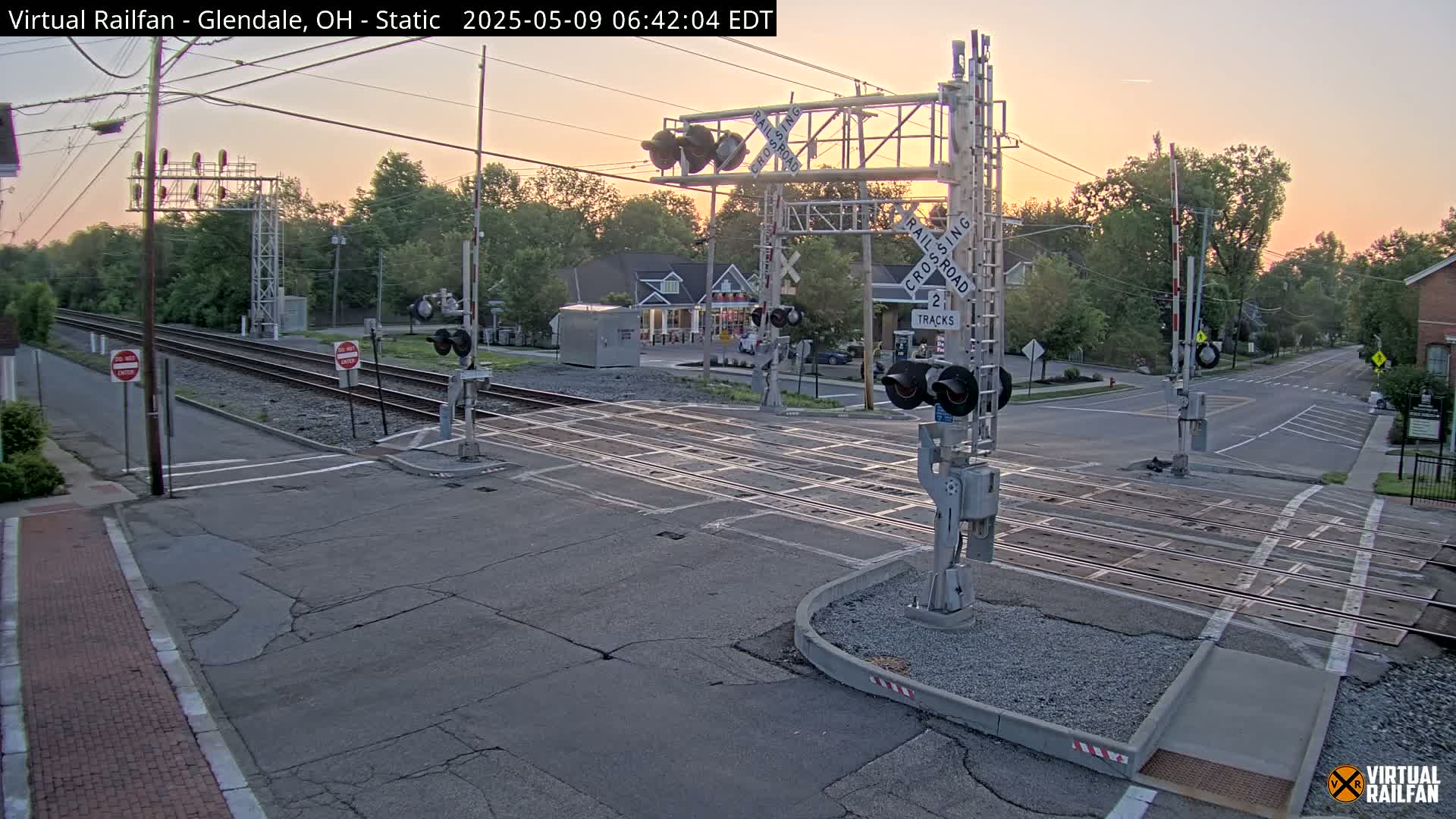 A railroad crossing with lowered gates is shown at sunrise, with two tracks intersecting a street in a residential area.