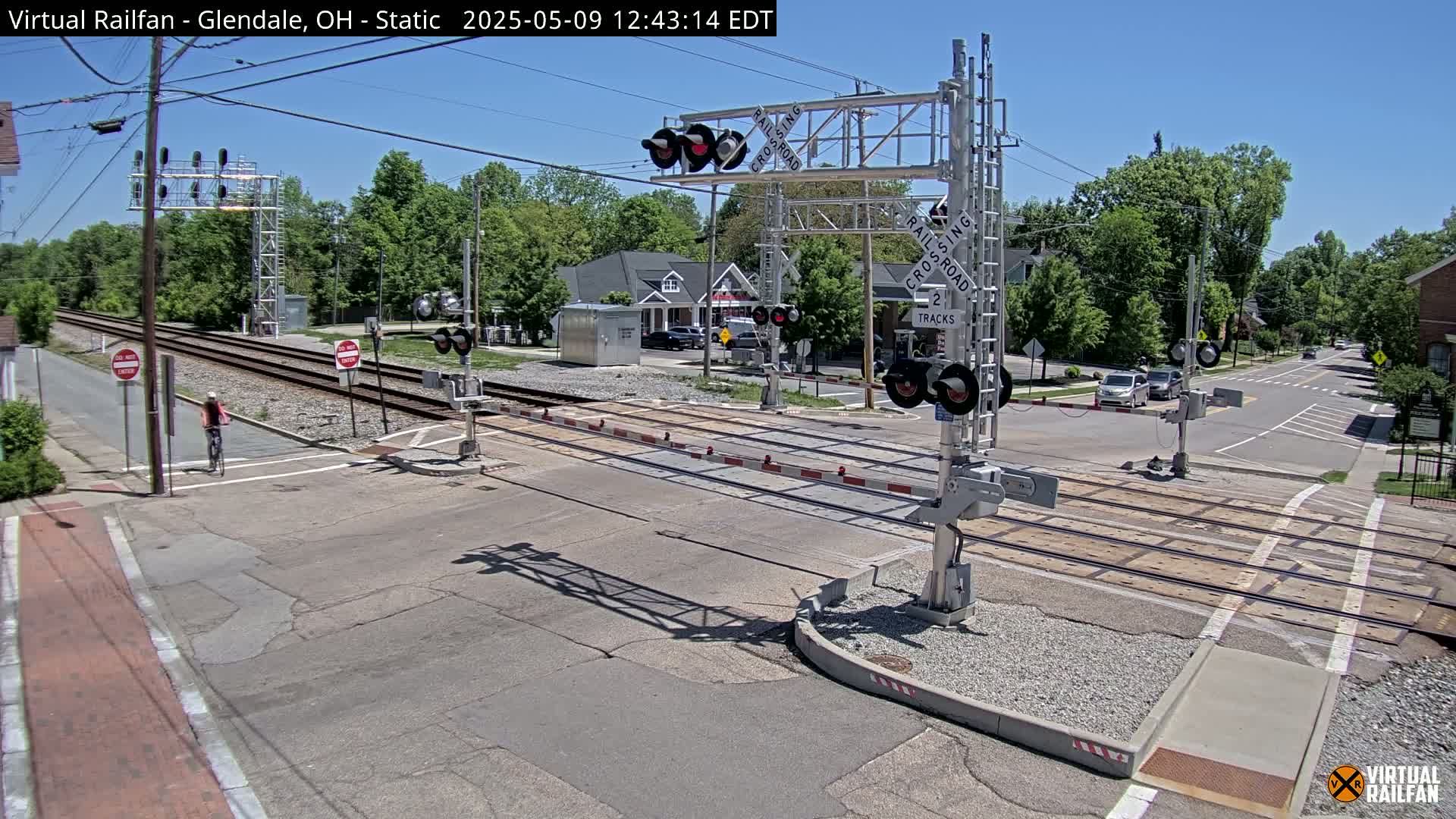 A railroad crossing with active signals and crossing gates under a sunny sky is seen from above.