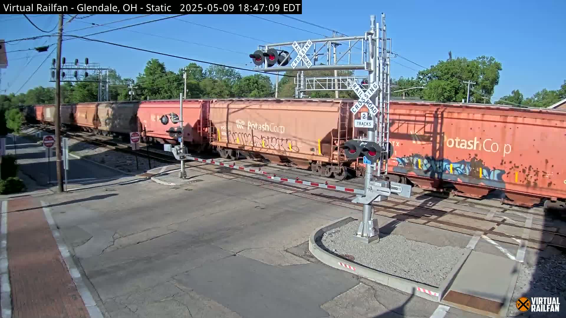 A long freight train, with several cars marked "PotashCorp," passes through a railroad crossing under a sunny sky.