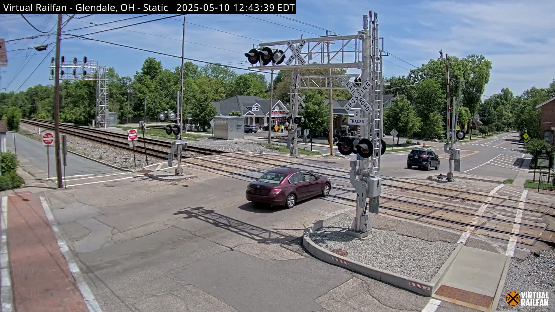 A maroon sedan is crossing a railroad intersection under a sunny sky, with trees and buildings visible in the background.
