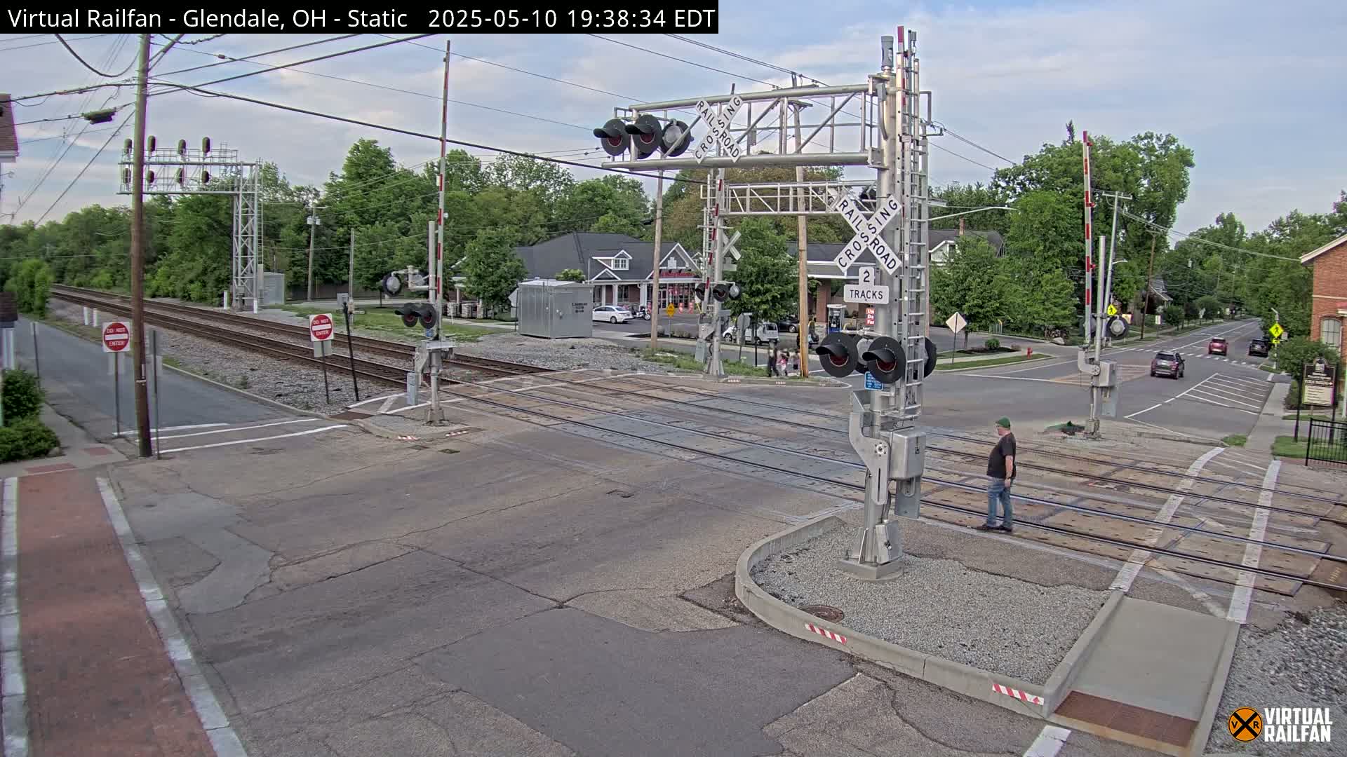 A mostly sunny view of a railroad crossing with a man walking near a train signal, several cars in the background, and lush green trees lining the streets.