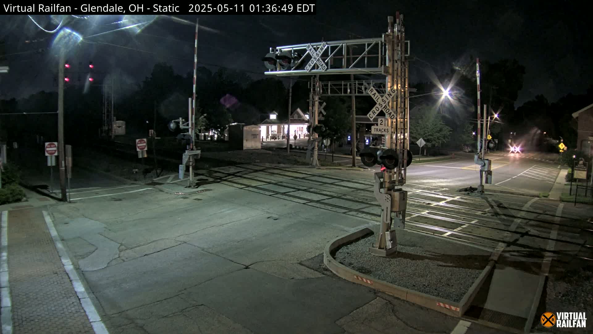 A nighttime view of a railroad crossing with lowered gates and flashing red lights, illuminated by streetlights and surrounded by dark trees.