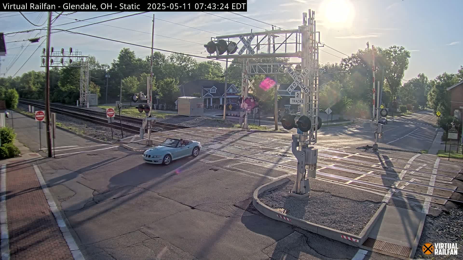 A light blue convertible car is crossing a sunny railroad intersection with its crossing gates down.
