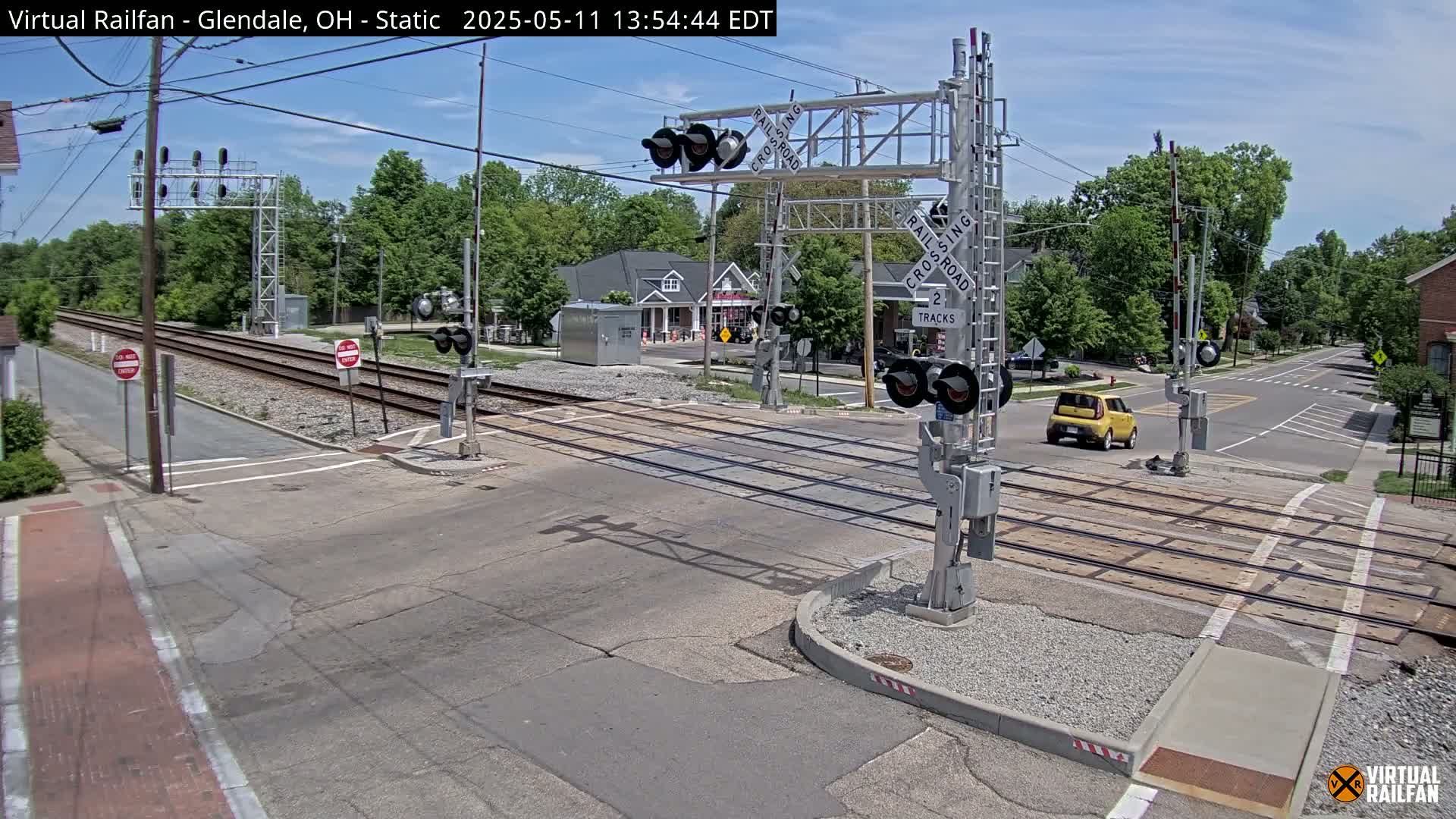 A street intersects a railroad crossing with two tracks under a sunny sky; a yellow car is stopped at the crossing.