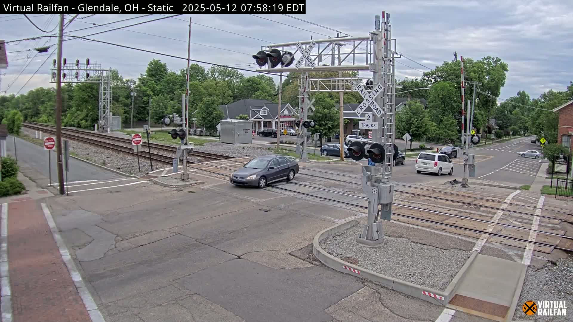 A gray sedan crosses a railroad intersection with lowered crossing gates under an overcast sky, while other vehicles wait nearby.