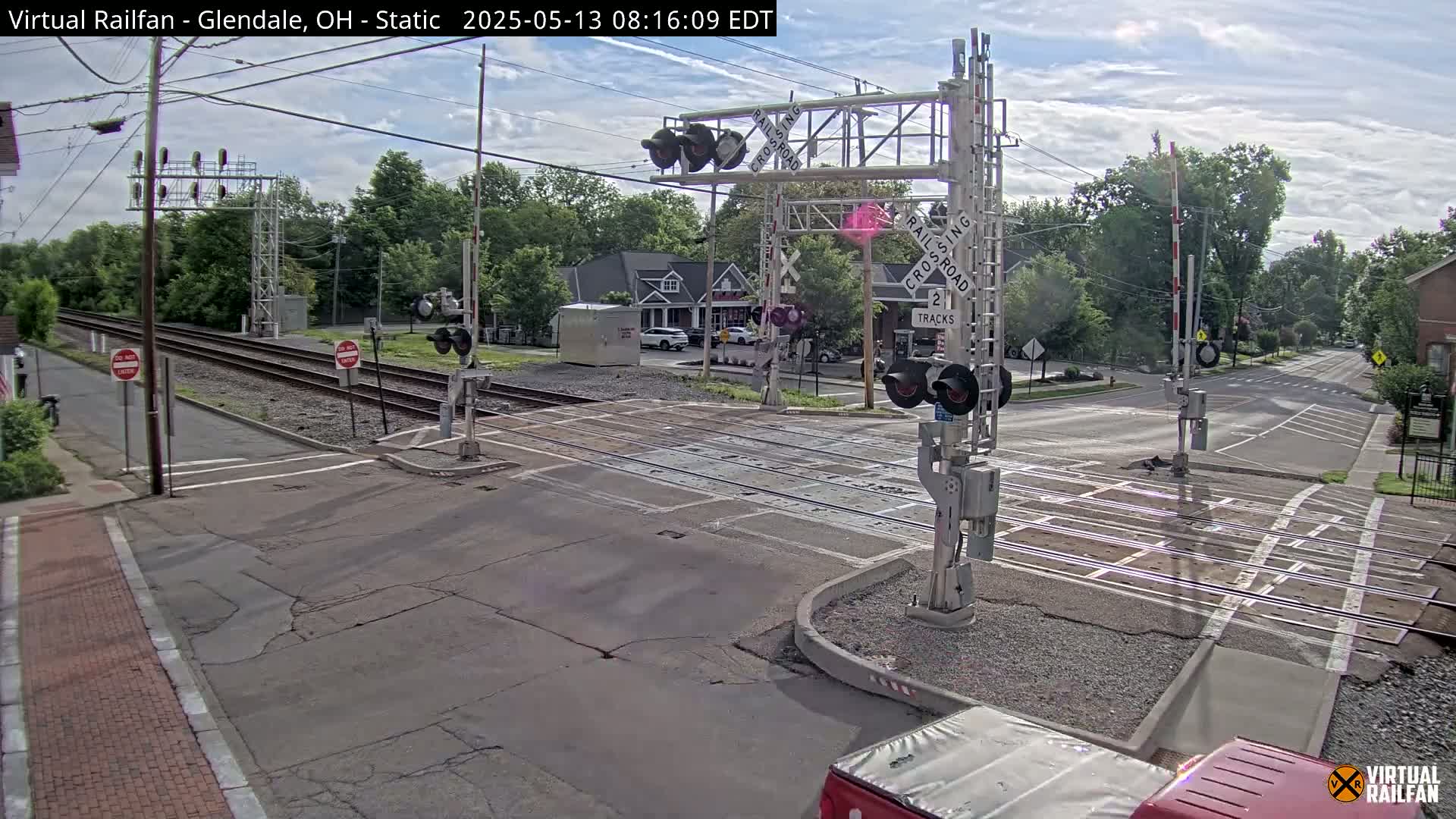 A mostly sunny day shows a railroad crossing with lowered gates and flashing lights, where two roads intersect the tracks.