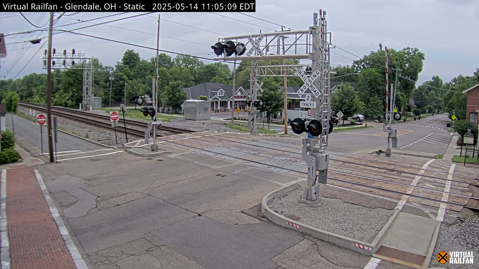 A railroad crossing with lowered gates is shown on an overcast day, with trees and buildings visible in the background.