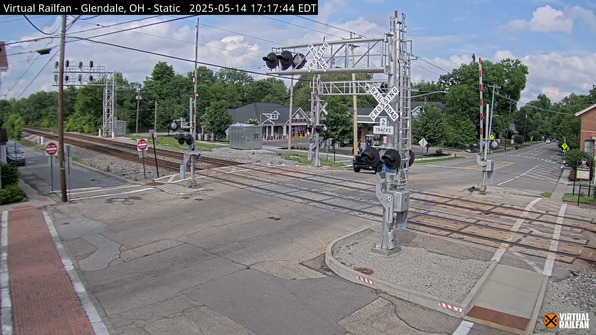 A railroad crossing with lowered gates and flashing lights on a sunny day, with a car stopped at the crossing and trees and buildings in the background.