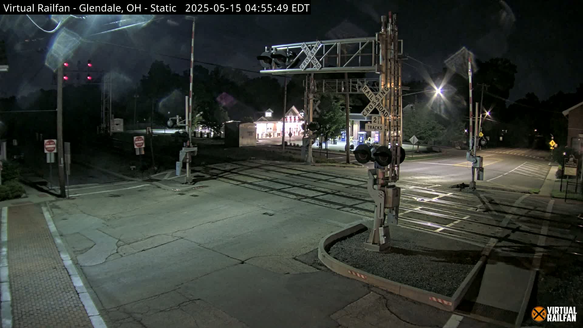 A nighttime view of a deserted railroad crossing with lowered gates, illuminated by streetlights and building lights.