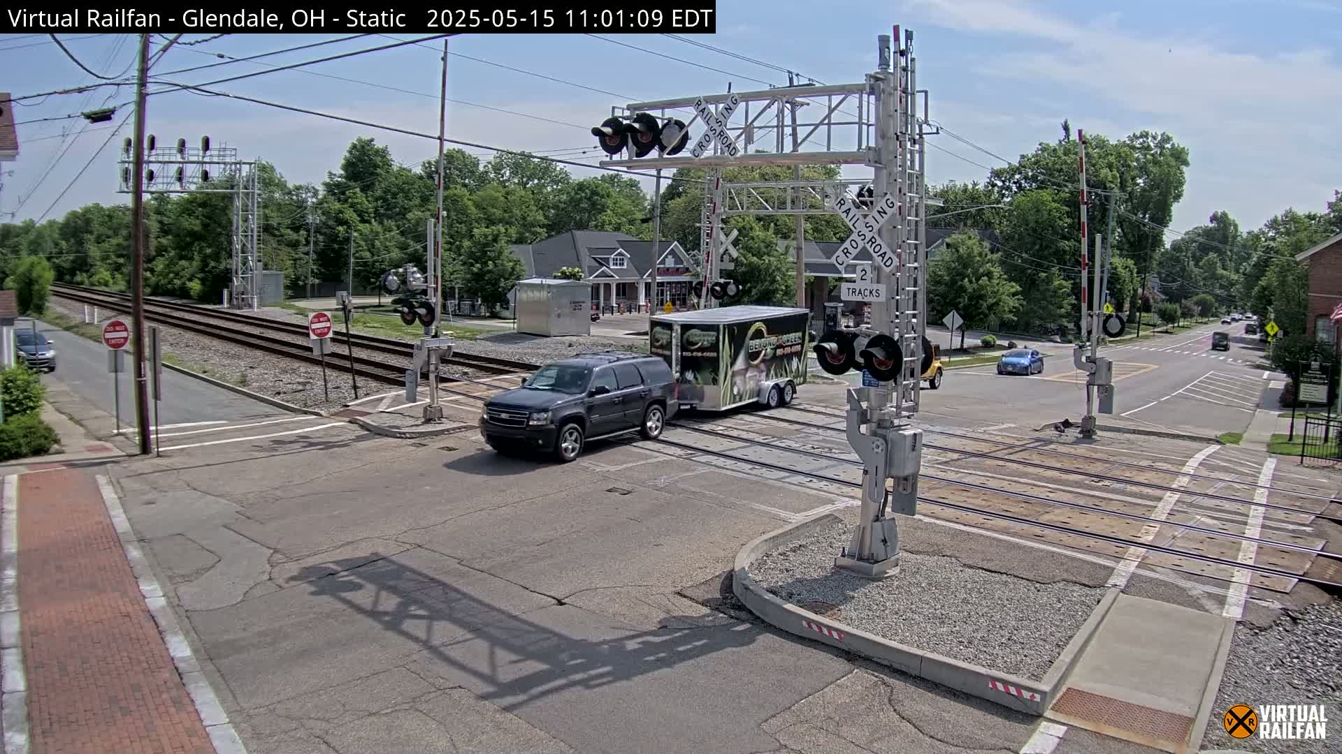 A dark-colored SUV towing a trailer is stopped at a railroad crossing on a sunny day.