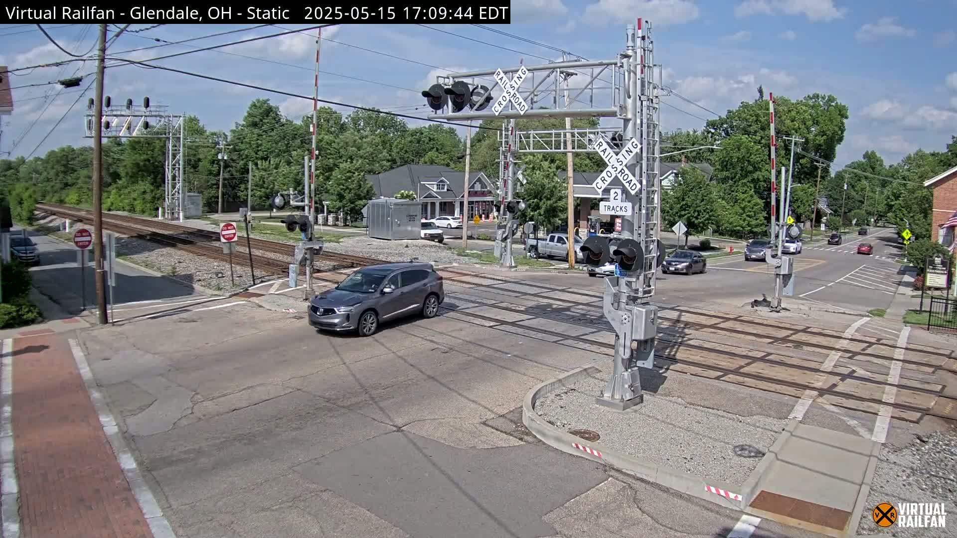 A gray SUV drives across a railroad crossing under a mostly sunny sky, with other vehicles at a nearby intersection.