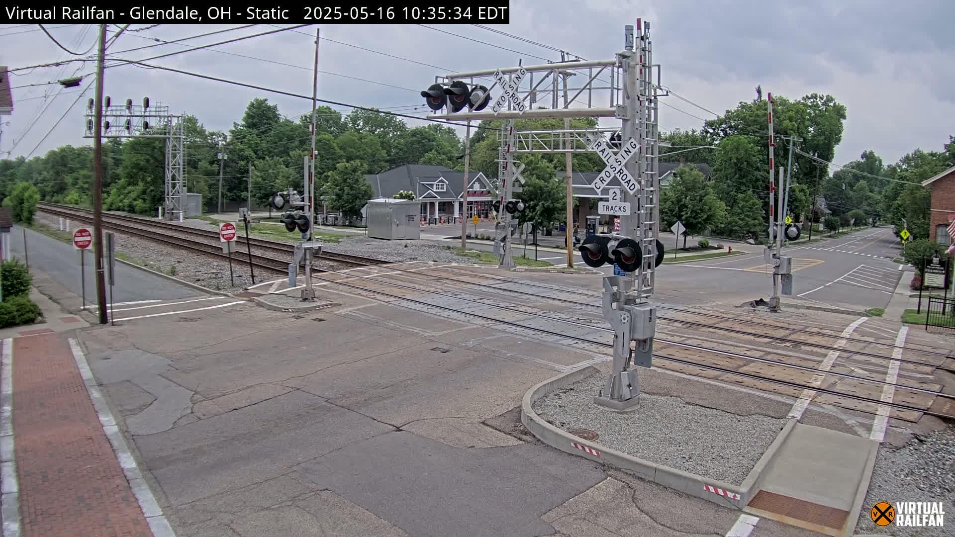 A railroad crossing with lowered gates is shown on an overcast day, with a street intersecting the tracks.
