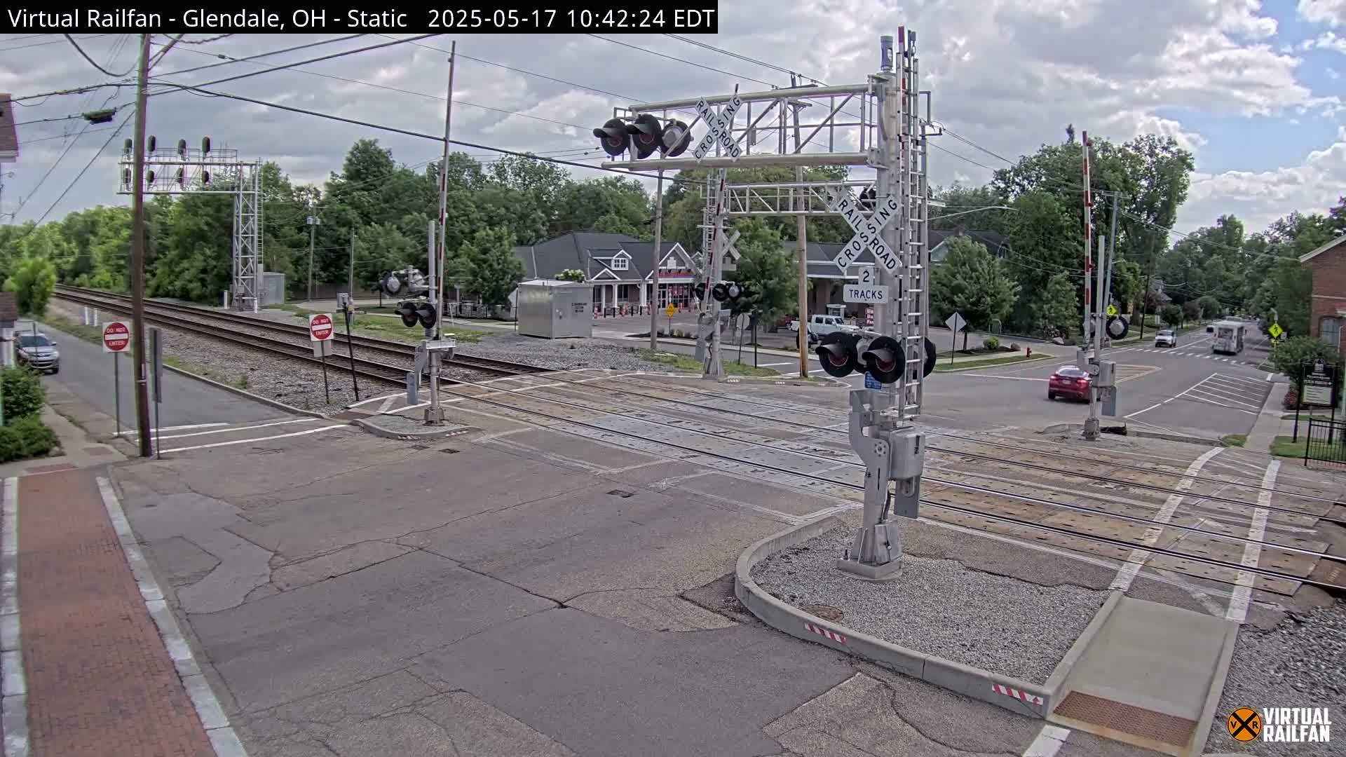 A partly cloudy day shows a railroad crossing with lowered gates, two tracks, and several cars at a street intersection near some buildings.