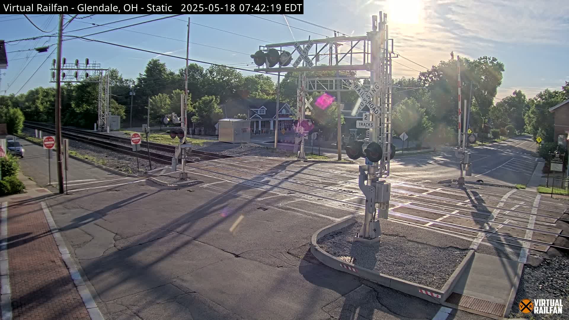 A sunny morning view of a railroad crossing with lowered gates, two tracks, and a road intersecting.