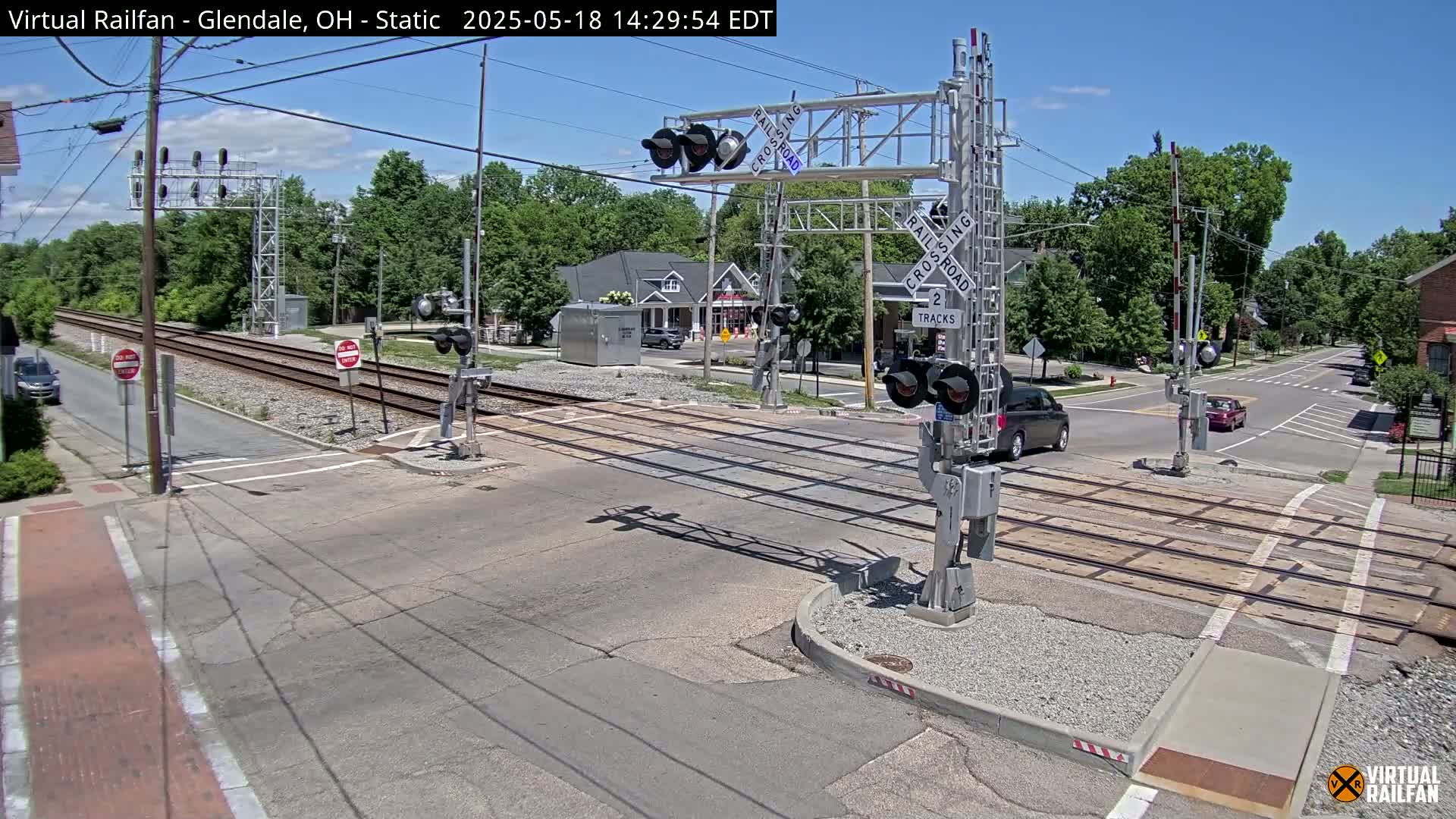 A sunny day at a railroad crossing where a road intersects with train tracks, showing a few cars and buildings in the background.