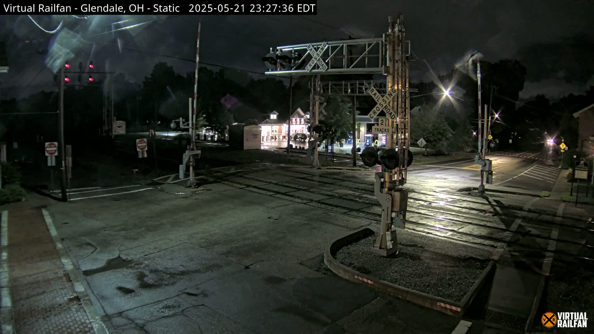 A nighttime view of a railroad crossing with lowered gates and flashing red lights, under partly cloudy skies.