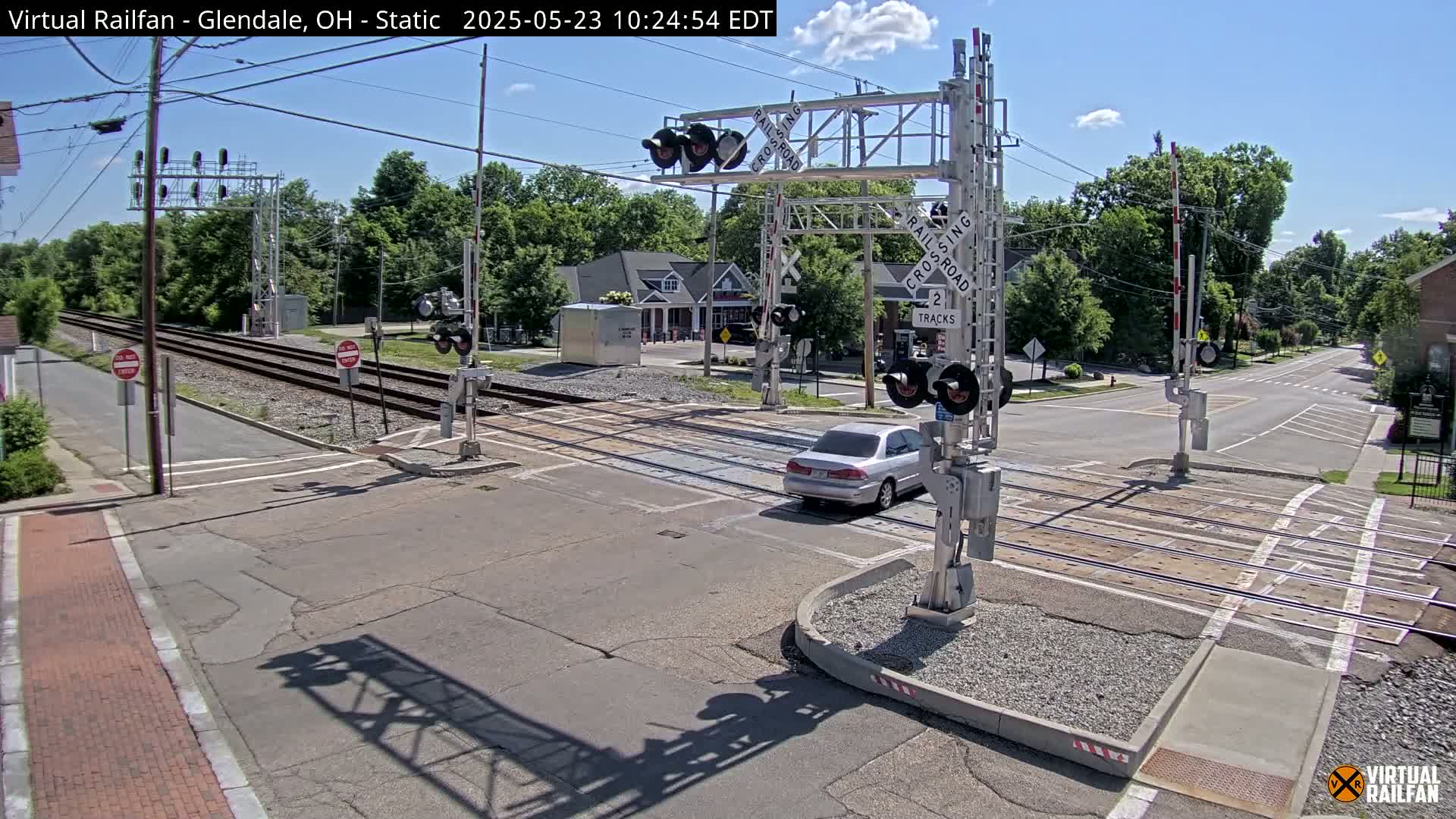 A silver car drives across a railroad crossing under a sunny sky, passing between two sets of crossing gates.