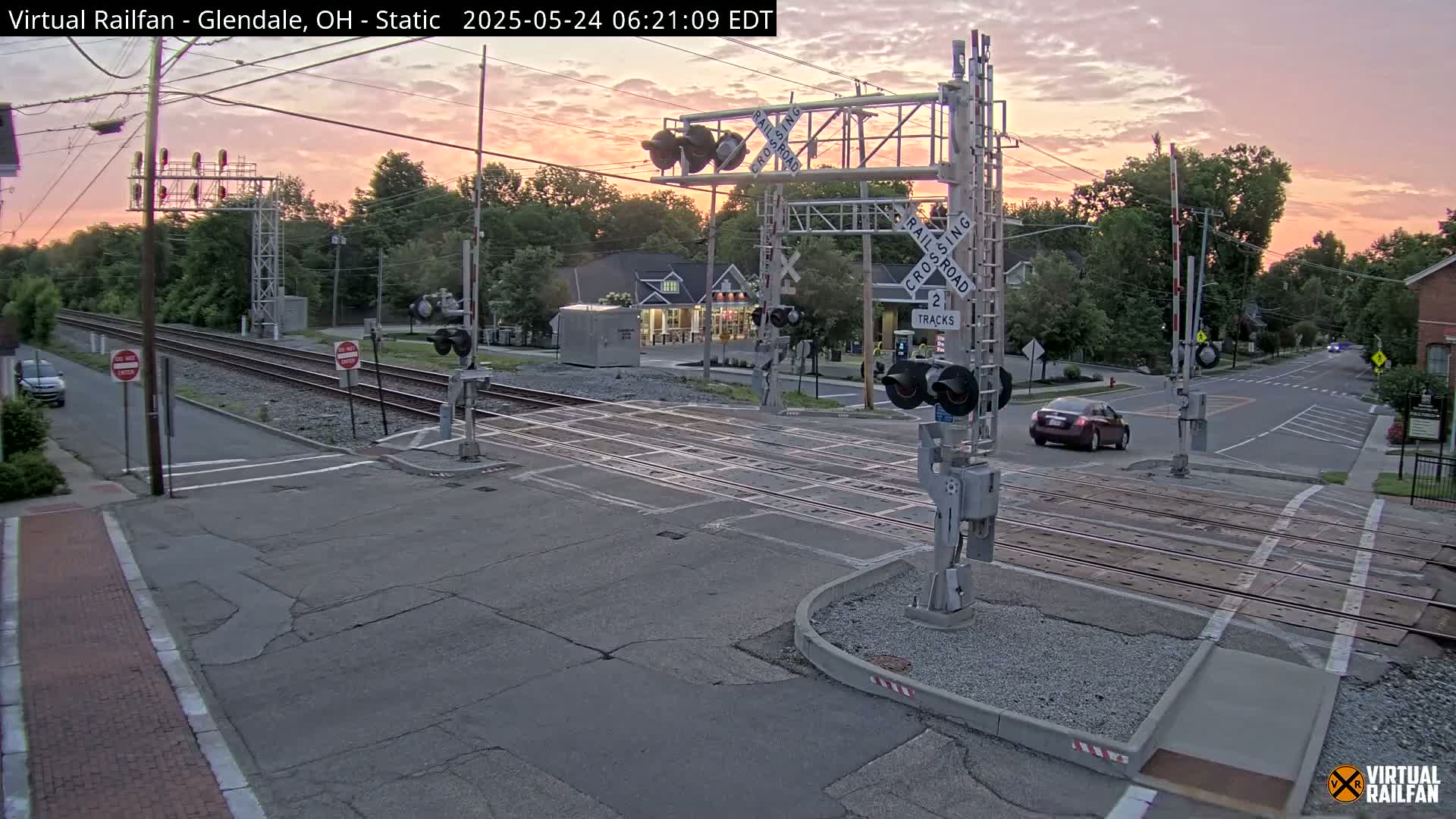 A railroad crossing with lowered gates is shown at sunrise, with one car stopped at the crossing and another driving away.