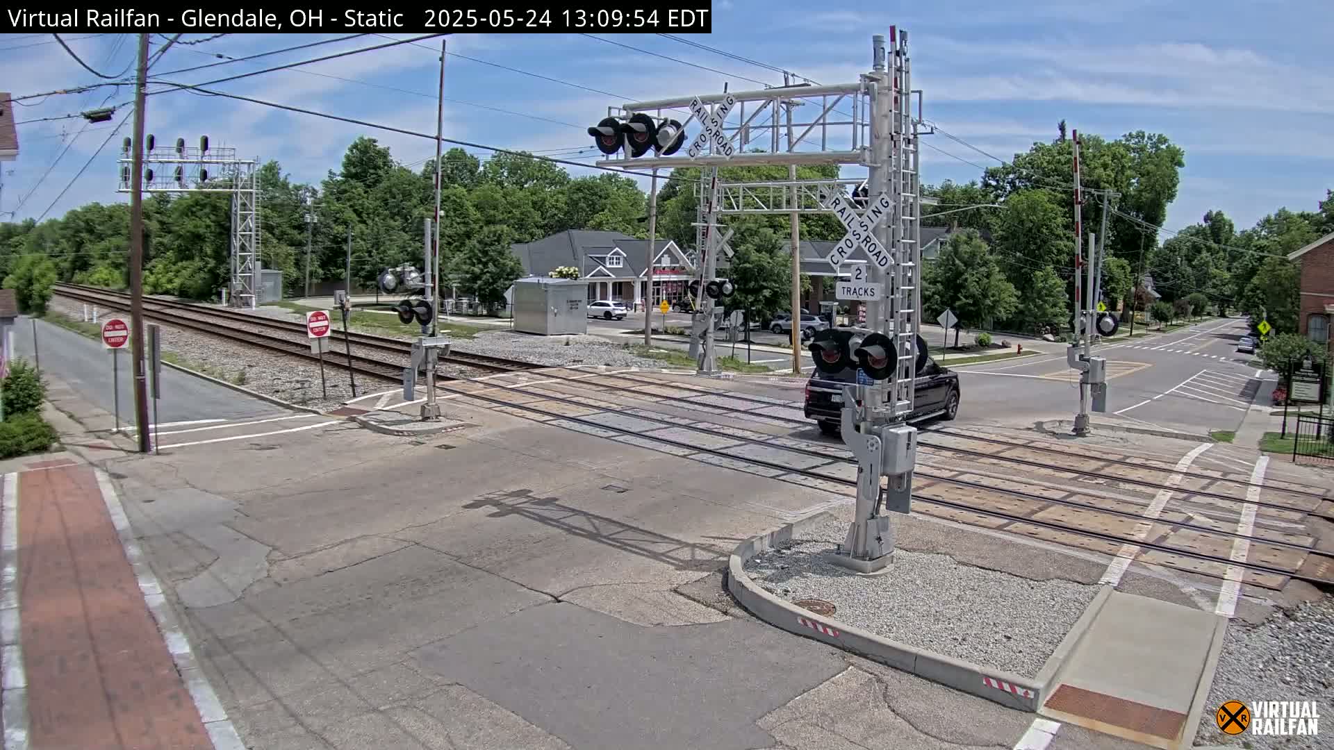 A sunny day shows a railroad crossing with a vehicle stopped, surrounded by residential buildings and trees.