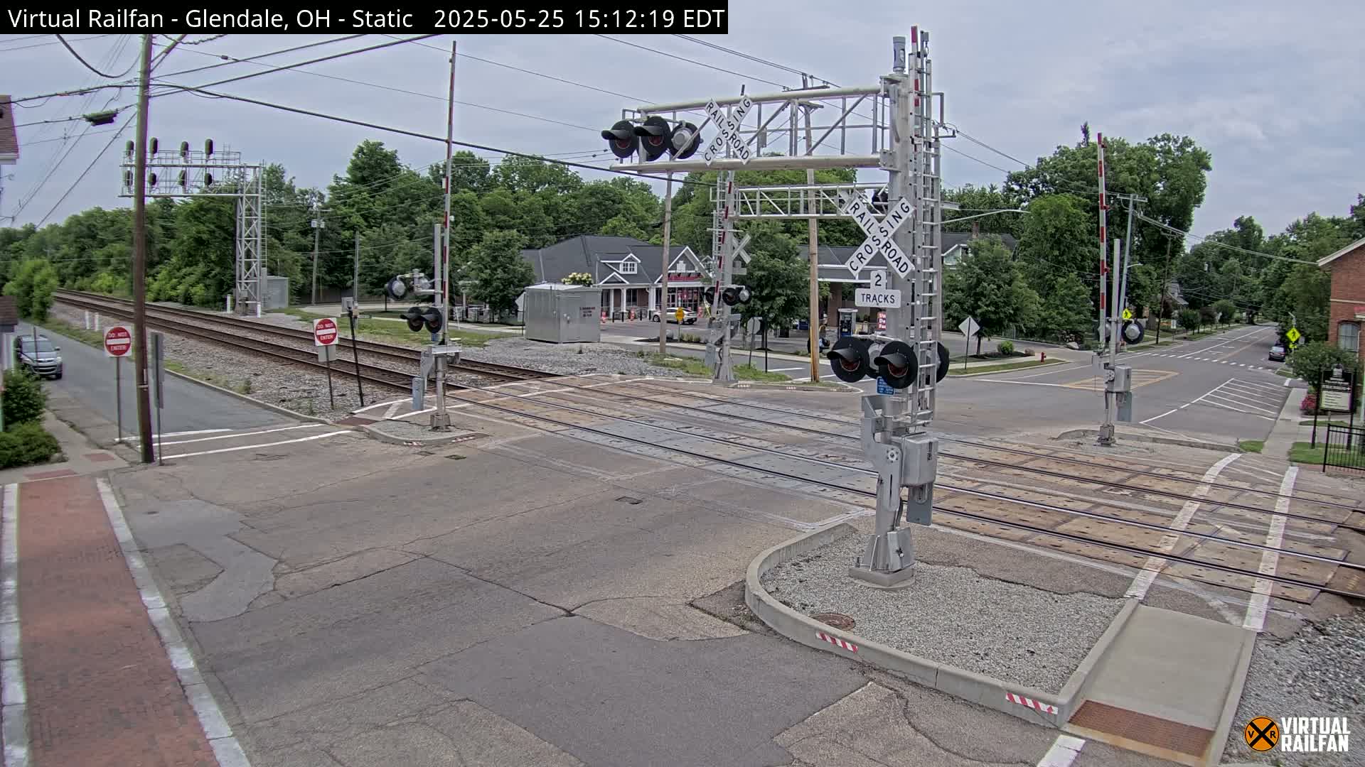 A railroad crossing with lowered gates is shown on an overcast day, with a road intersecting the train tracks and cars visible in the distance.