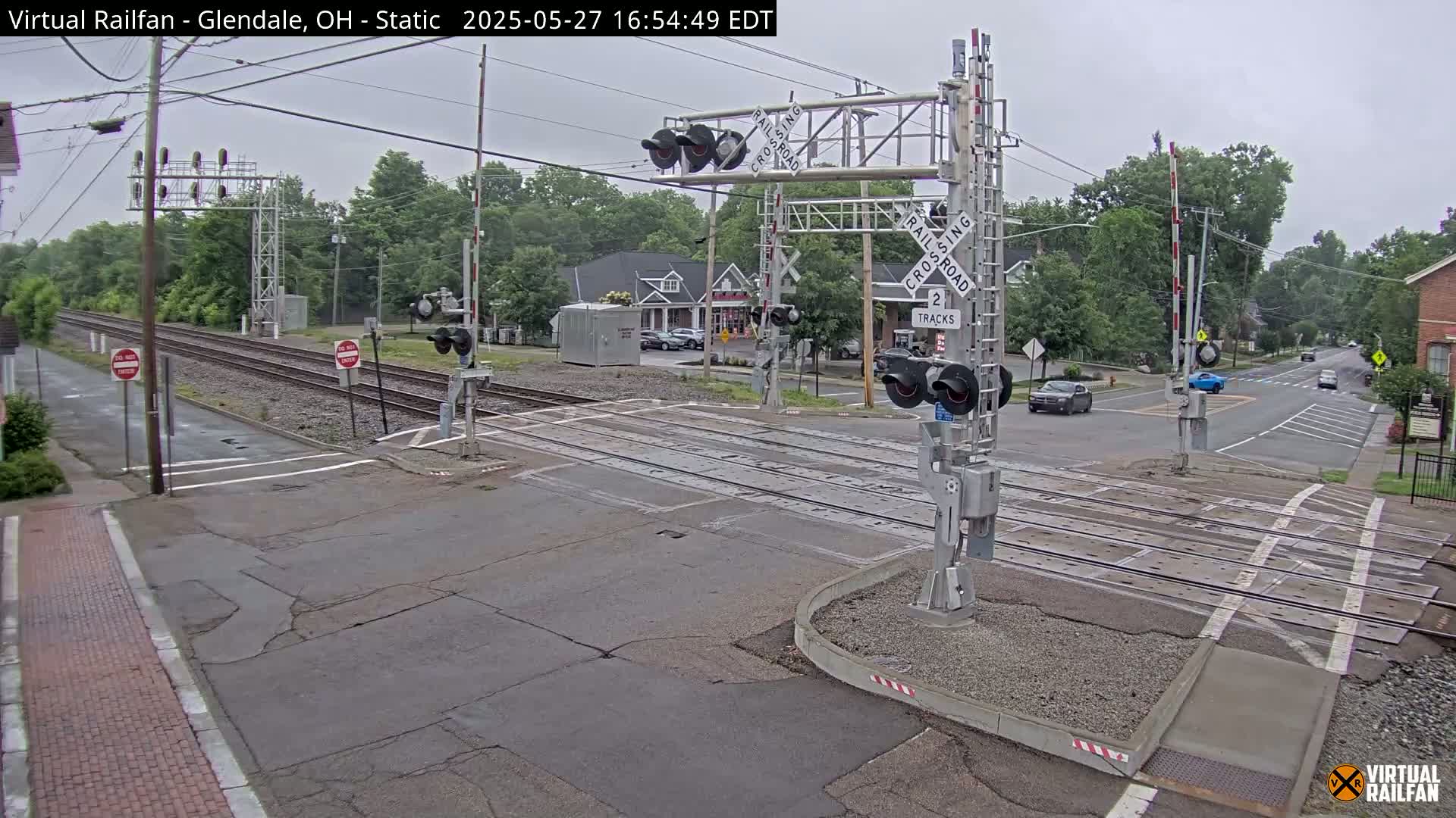 A railroad crossing with lowered gates shows two tracks intersecting a road under an overcast sky.