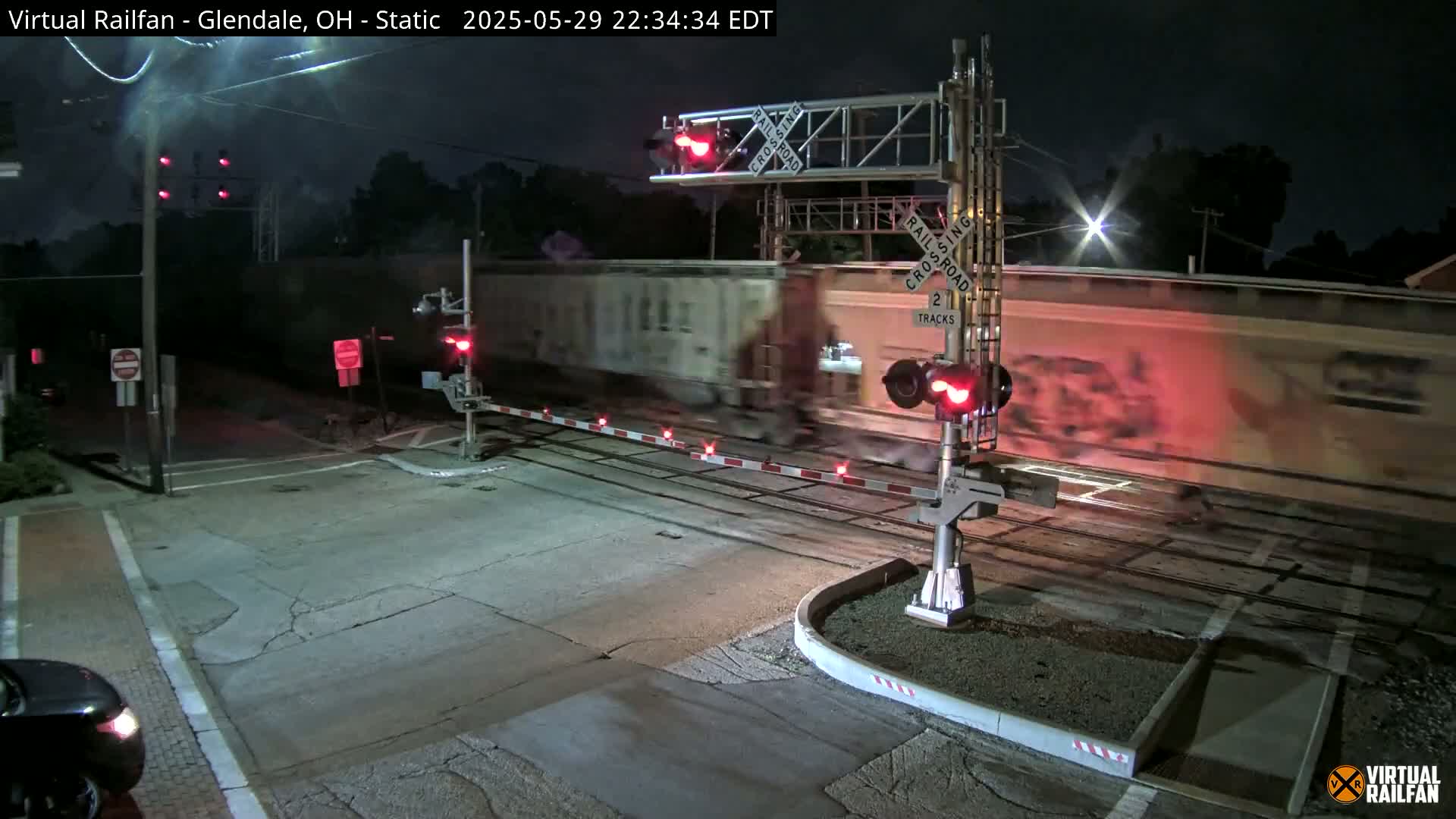 A long freight train passes through a railroad crossing at night, with the crossing gates down and lights flashing.