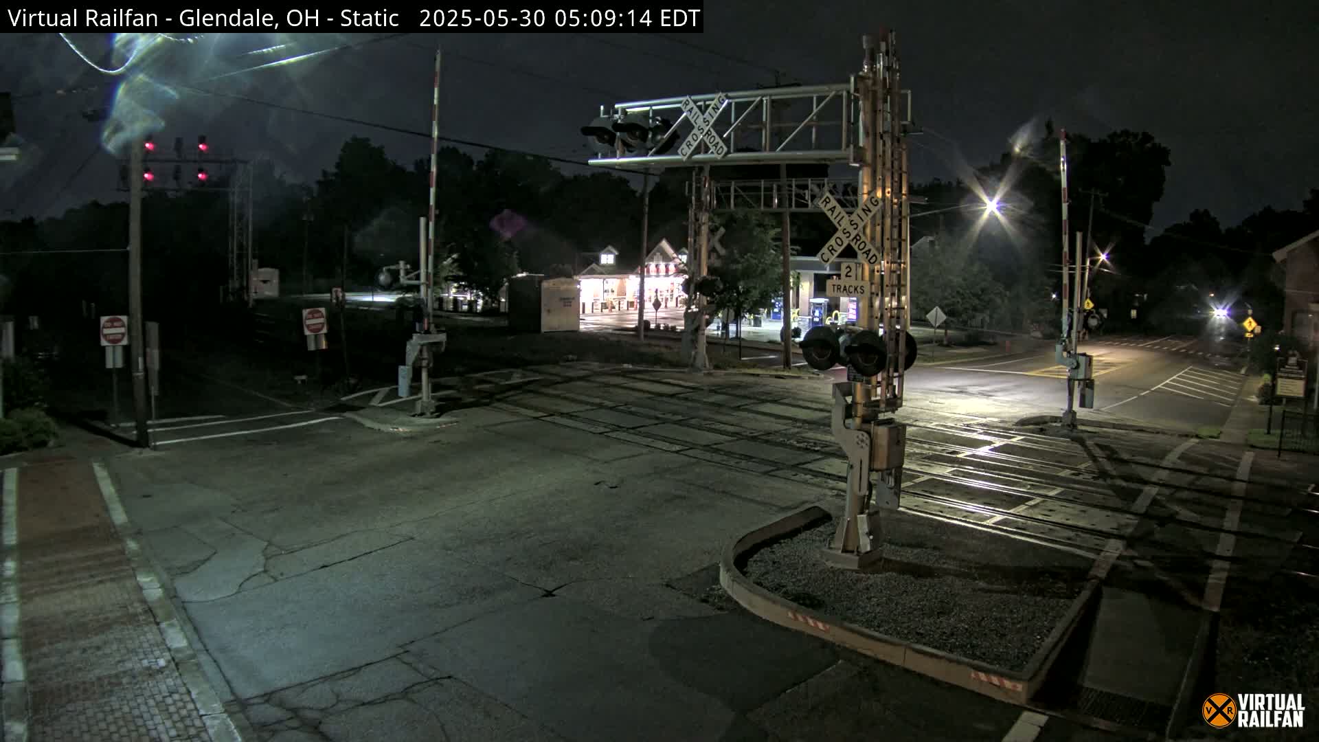 A nighttime view of a railroad crossing with lowered gates, illuminated by streetlights and the glow of nearby buildings.