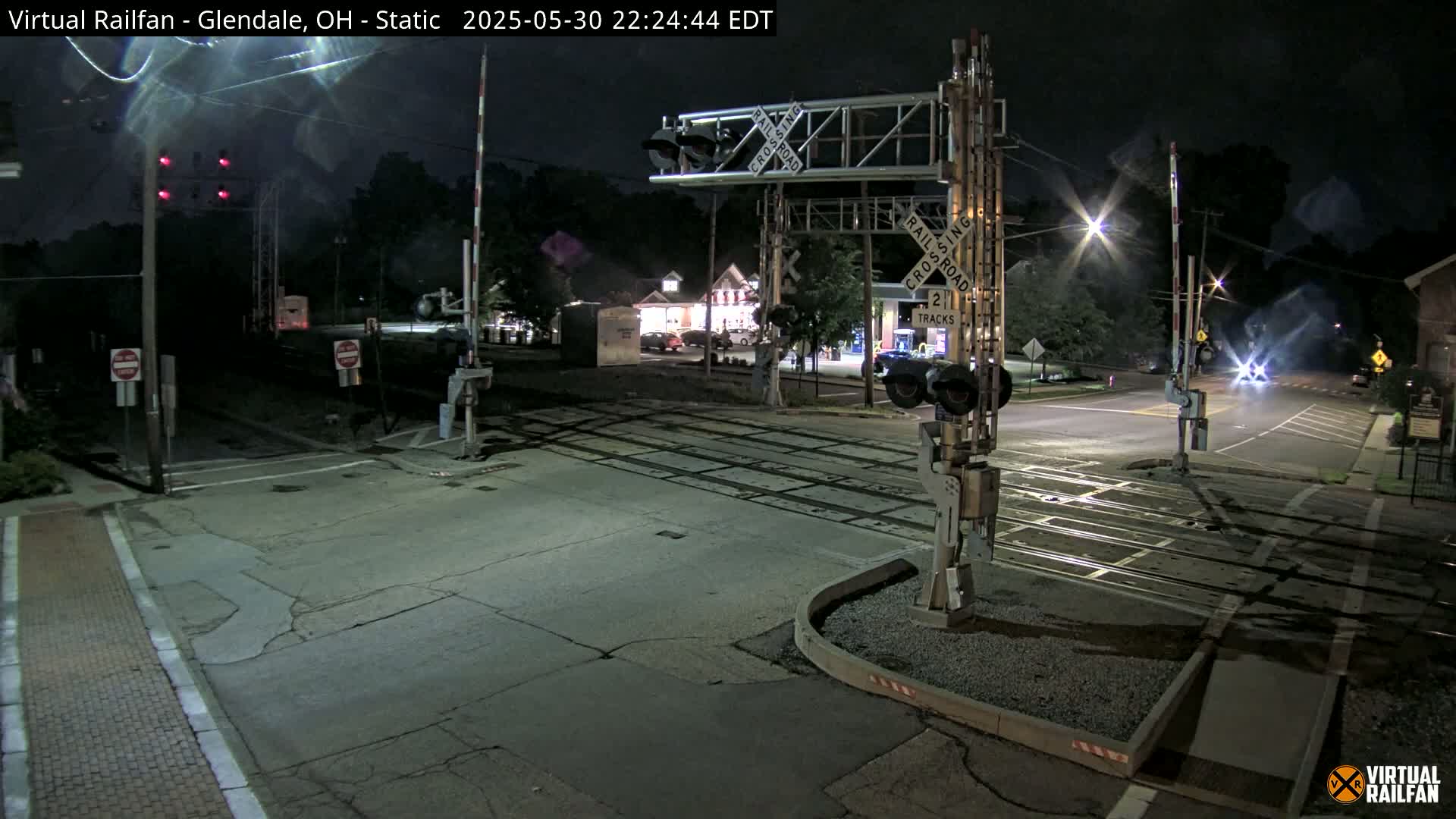 A nighttime view of a railroad crossing with lowered gates, showing a street intersection and some buildings in the background under clear skies.