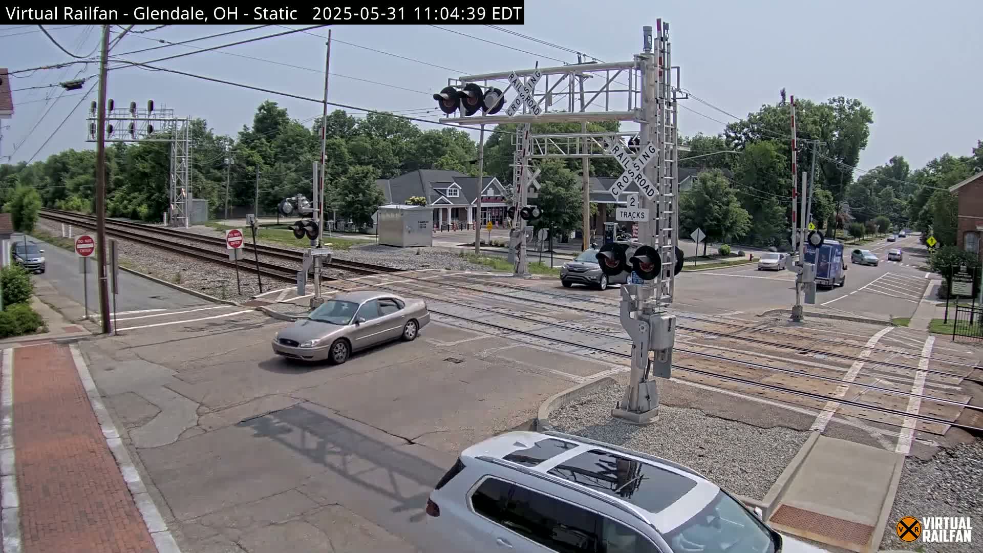 A mostly sunny day shows a street intersection with railroad tracks, crossing gates, and several cars; some are stopped and others are moving.