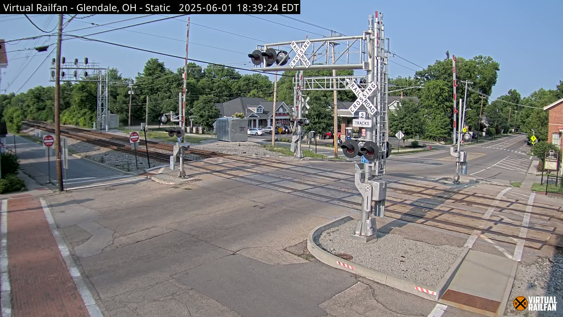 A railroad crossing with lowered gates is situated at a street intersection on a sunny day.