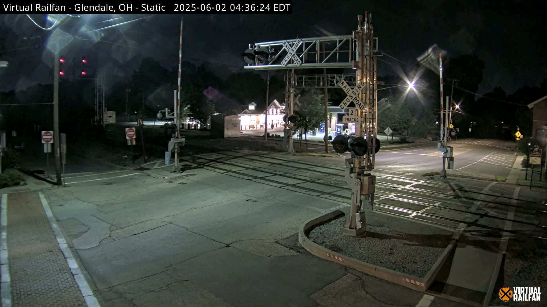 A nighttime view of a railroad crossing with lowered gates and flashing lights, illuminated by streetlights.