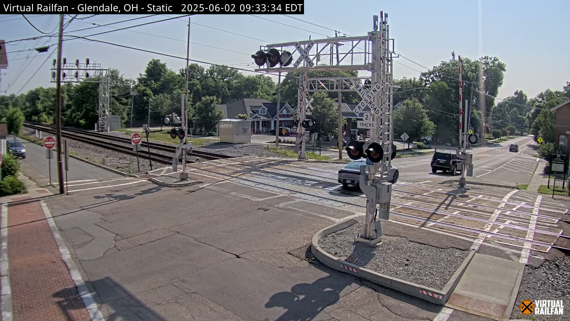 A sunny day shows a railroad crossing with lowered gates and a few vehicles waiting near the intersection.