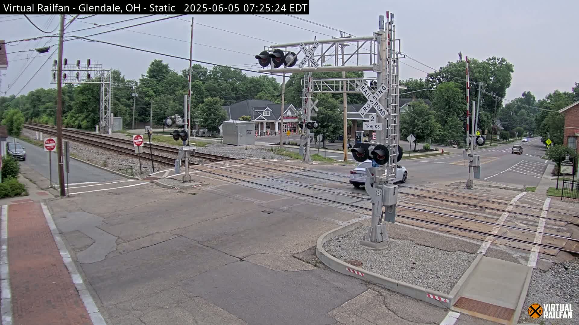 A railroad crossing with lowered gates and a car waiting at a road intersection under an overcast sky.