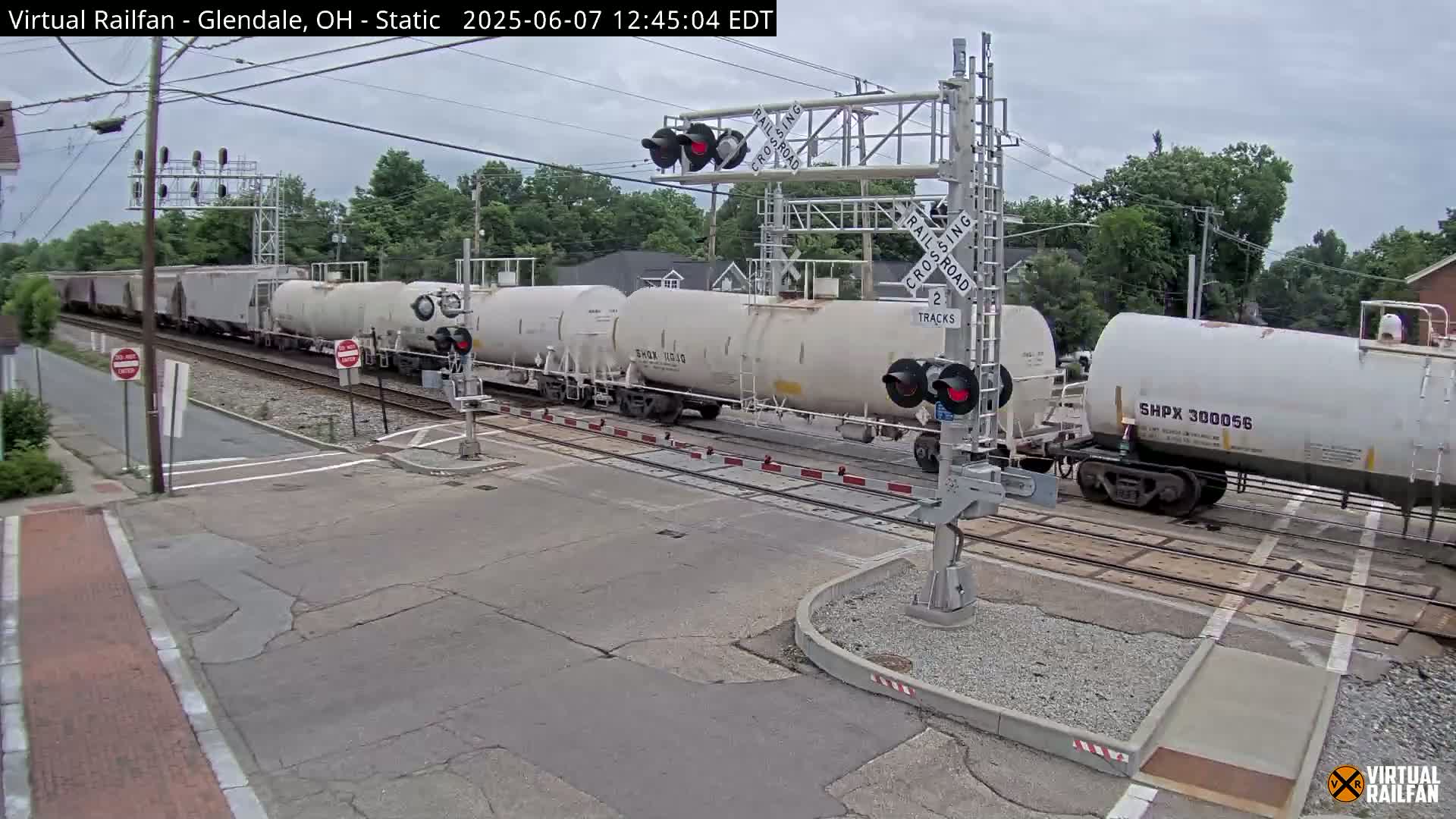A long freight train of white tanker cars passes through a railroad crossing on an overcast day.