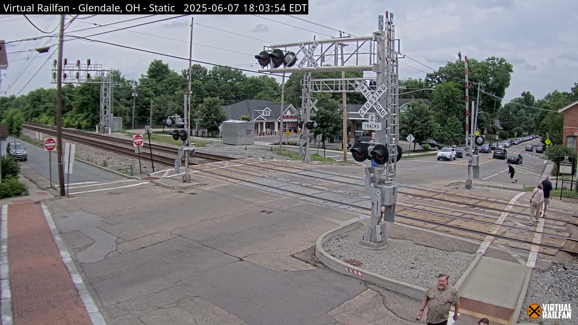 A railroad crossing with lowered gates is shown on an overcast day, with several cars stopped on the road and pedestrians crossing nearby.