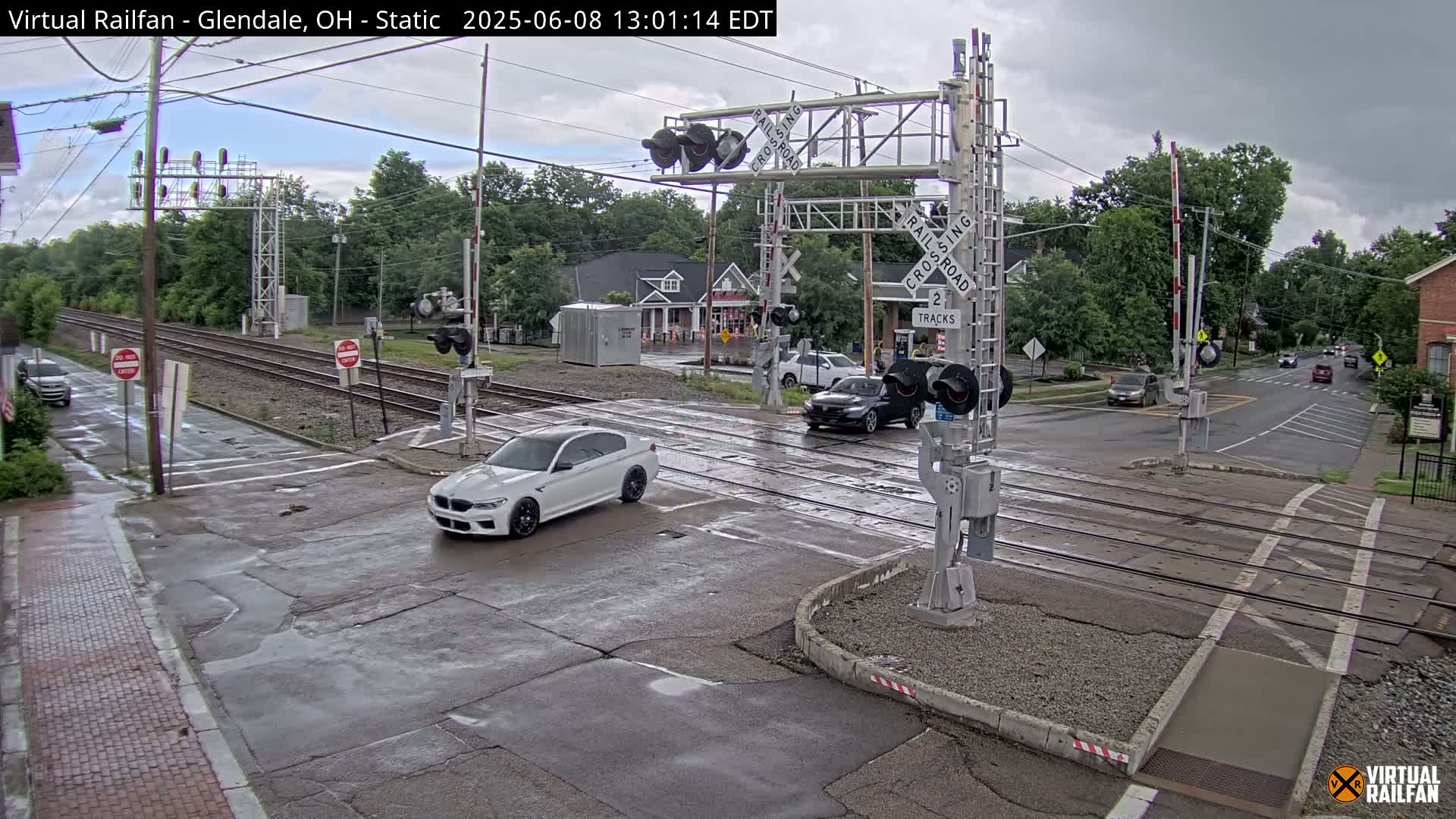 A wet road intersects a railroad crossing with several cars present under an overcast sky.