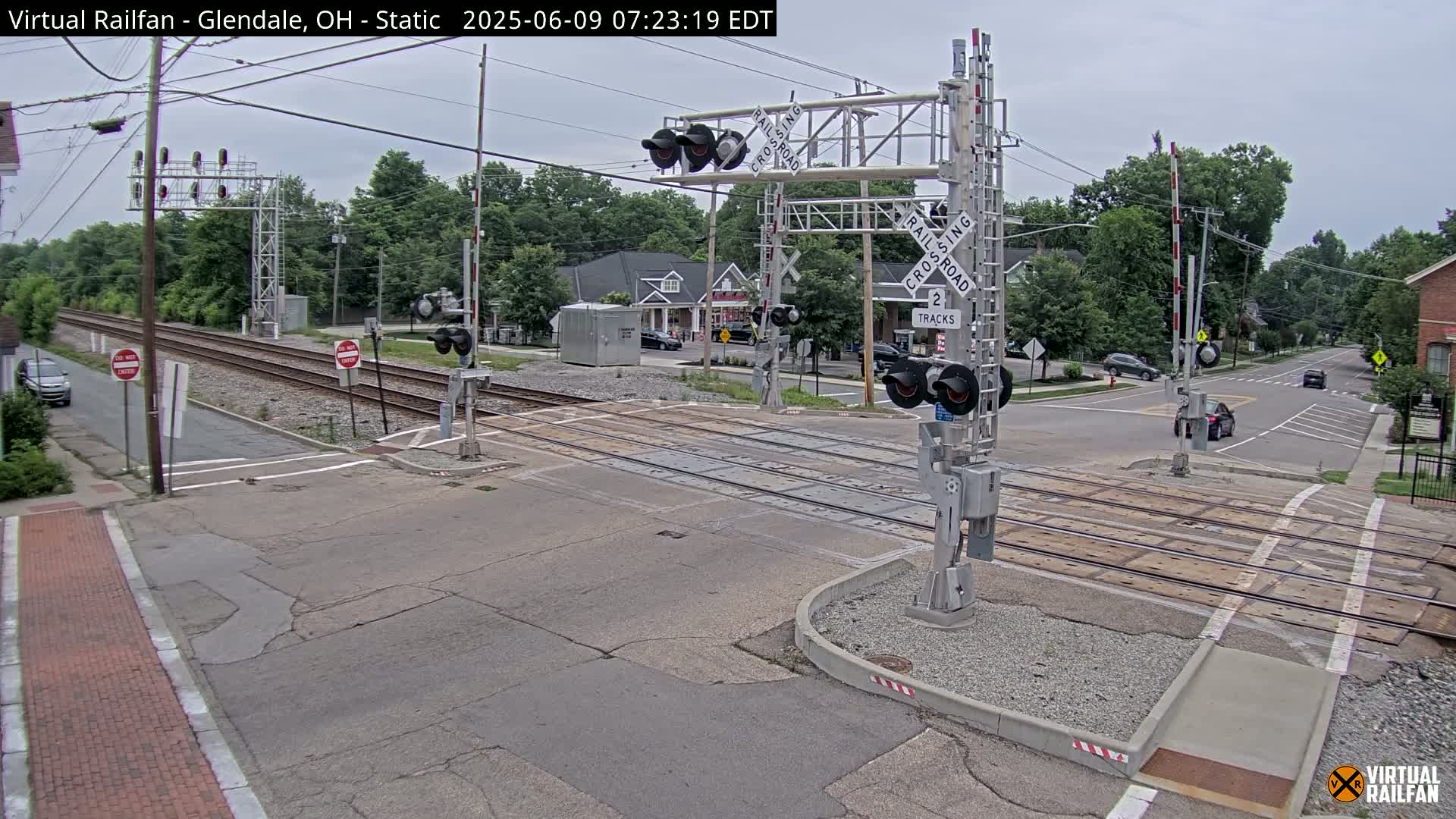 A railroad crossing with lowered gates and warning lights is shown on an overcast day, with several cars visible on the surrounding roads.