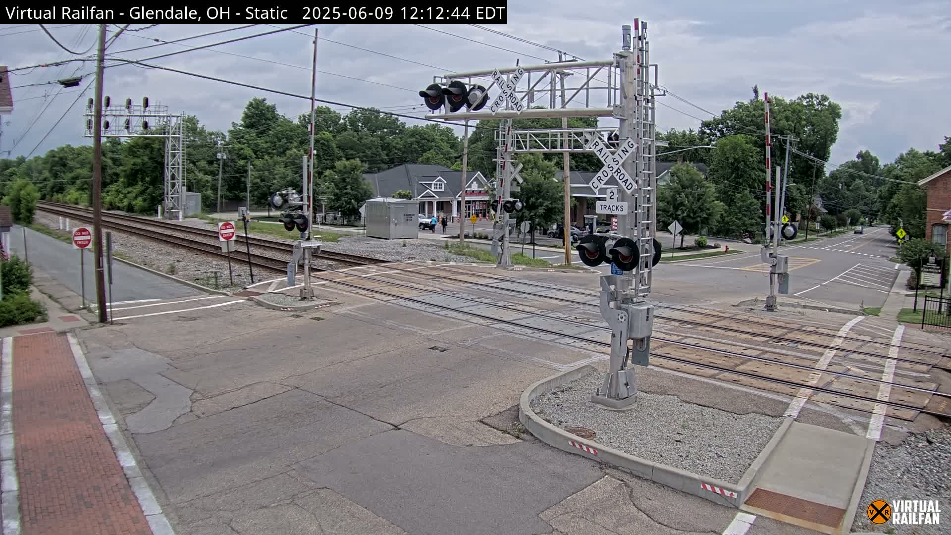 A railroad crossing with lowered gates is shown on an overcast day, with a road intersecting the tracks and buildings visible in the background.