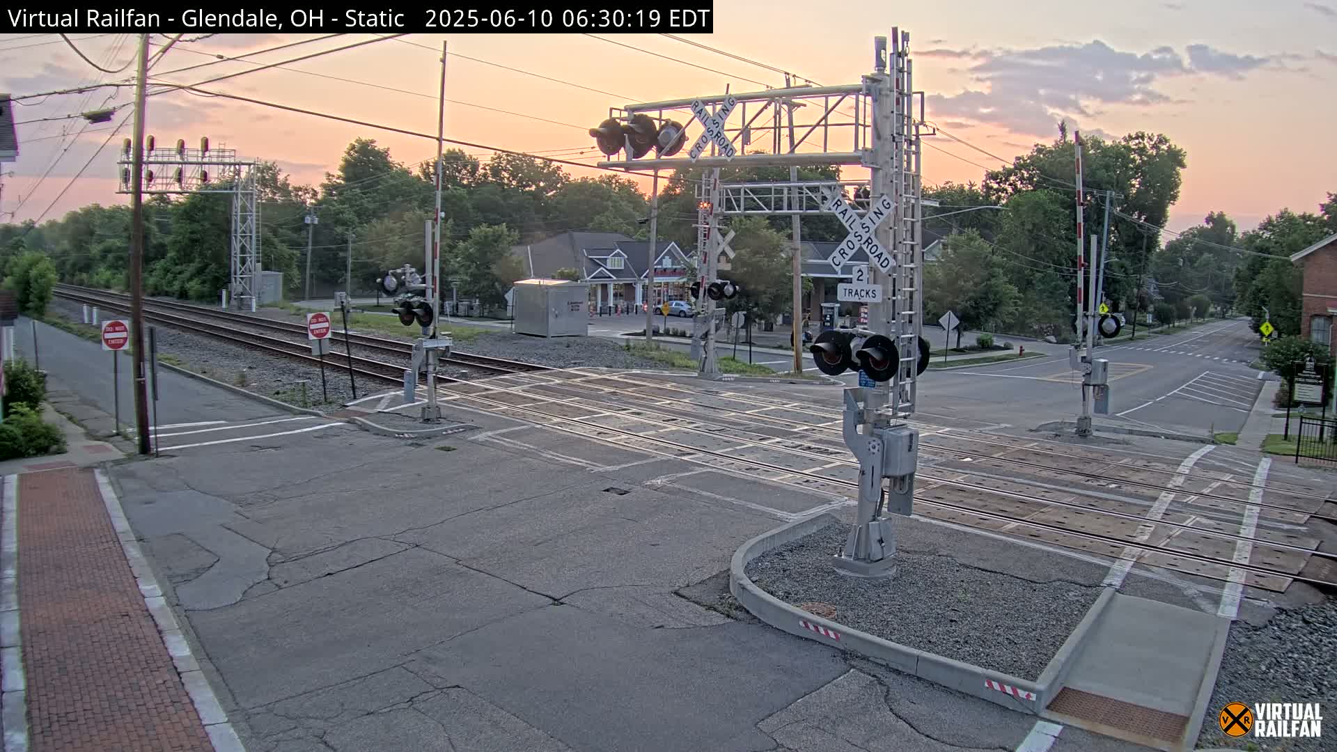 A railroad crossing with lowered gates is shown at sunrise, under a mostly clear sky.