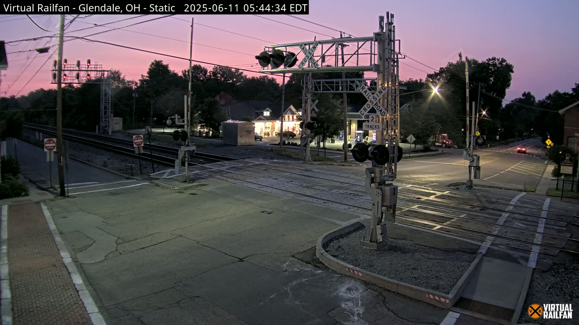 A railroad crossing at twilight shows lowered gates and flashing lights, with a road intersecting the tracks under a dusky pink sky.