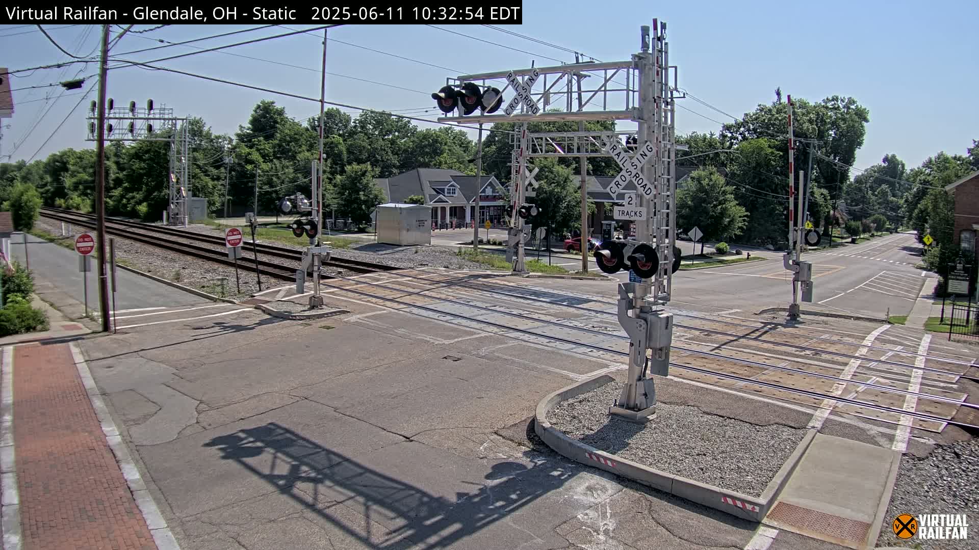 A sunny day at a railroad crossing shows two sets of tracks intersecting a road, with crossing gates and signals in place.