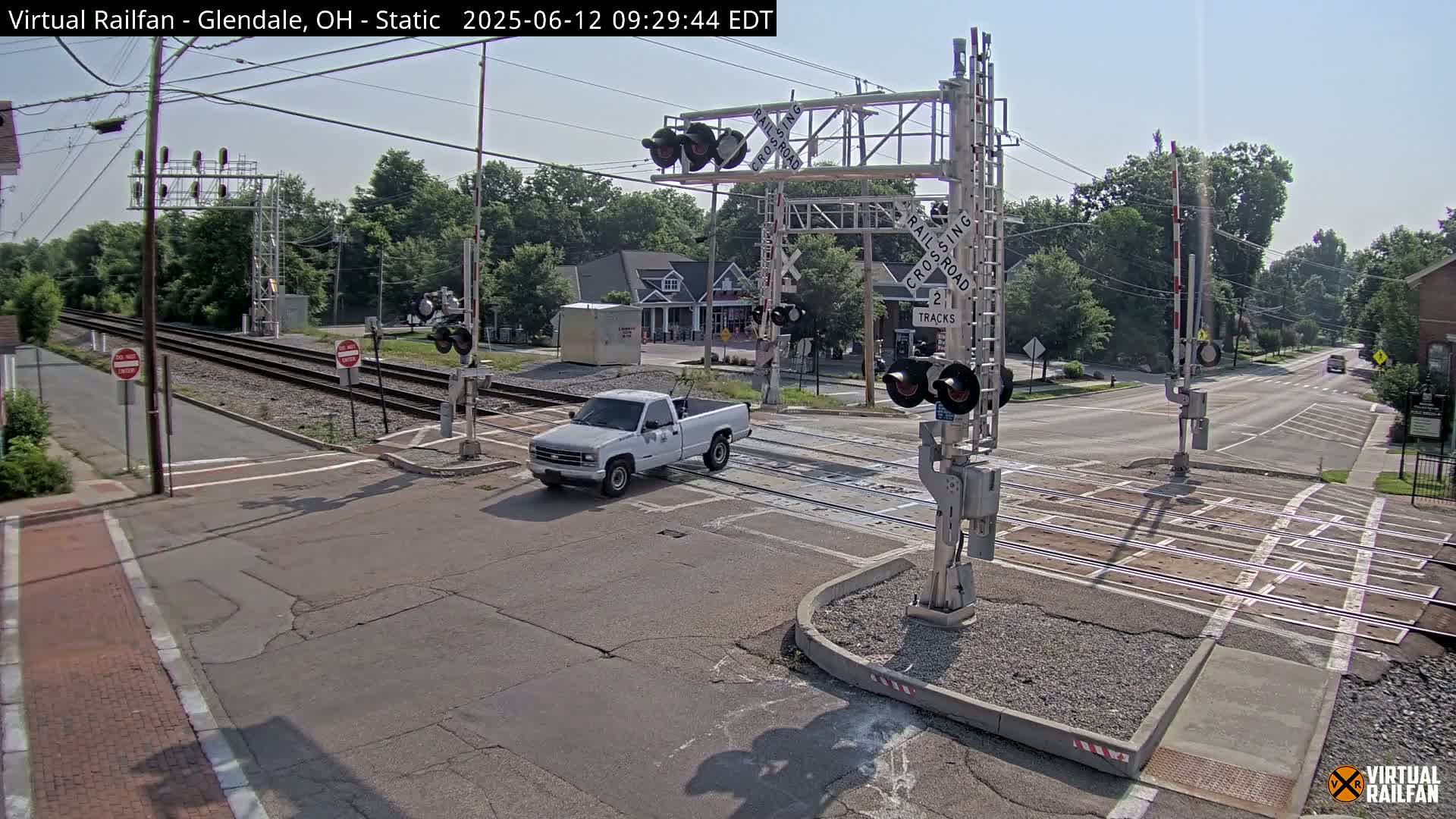 A white pickup truck is crossing a railroad intersection under a sunny sky, with railroad crossing gates and signals in place.