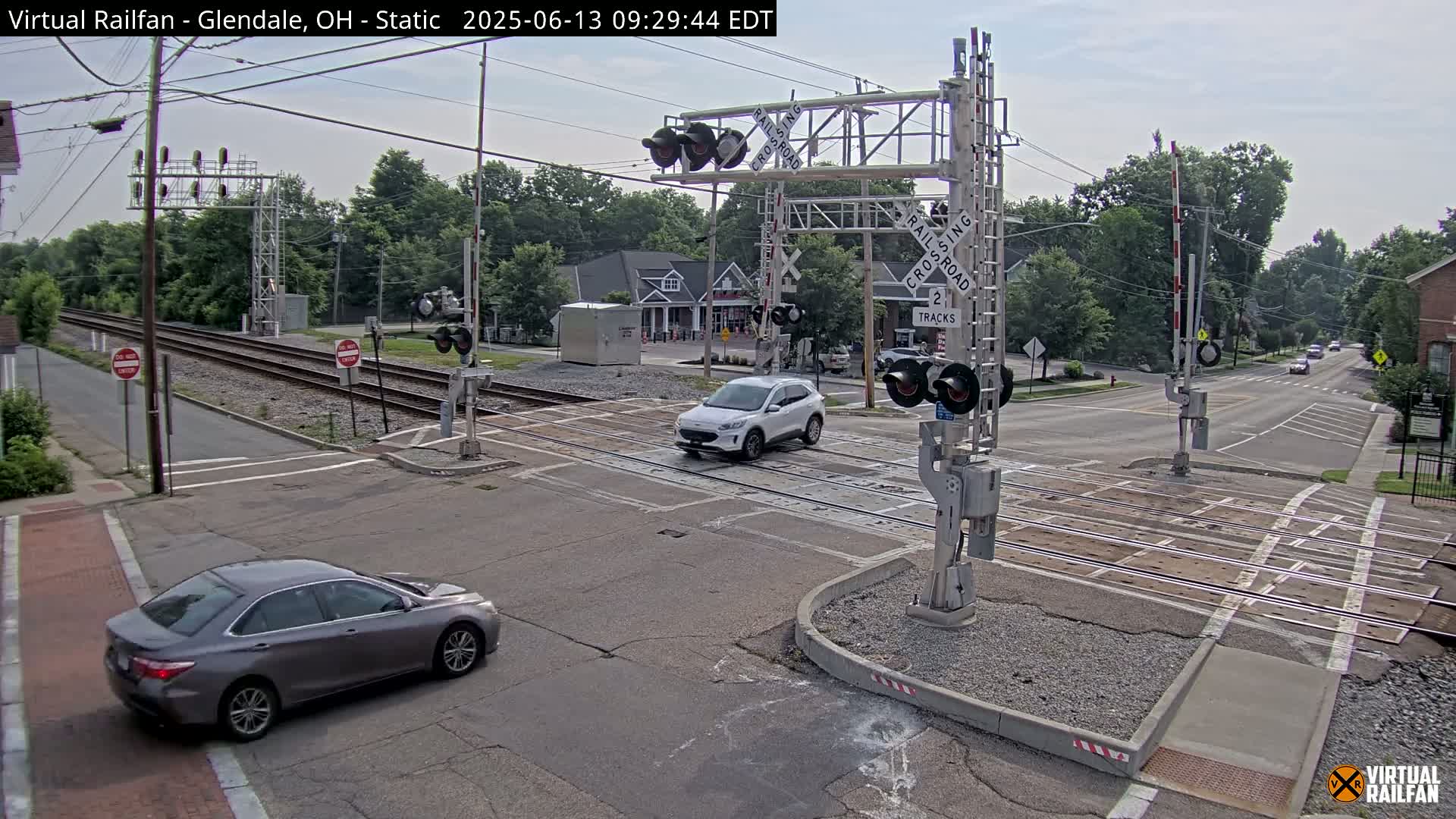 A mostly sunny day shows a street intersection with railroad tracks, crossing gates, and several cars, one of which is crossing the tracks.