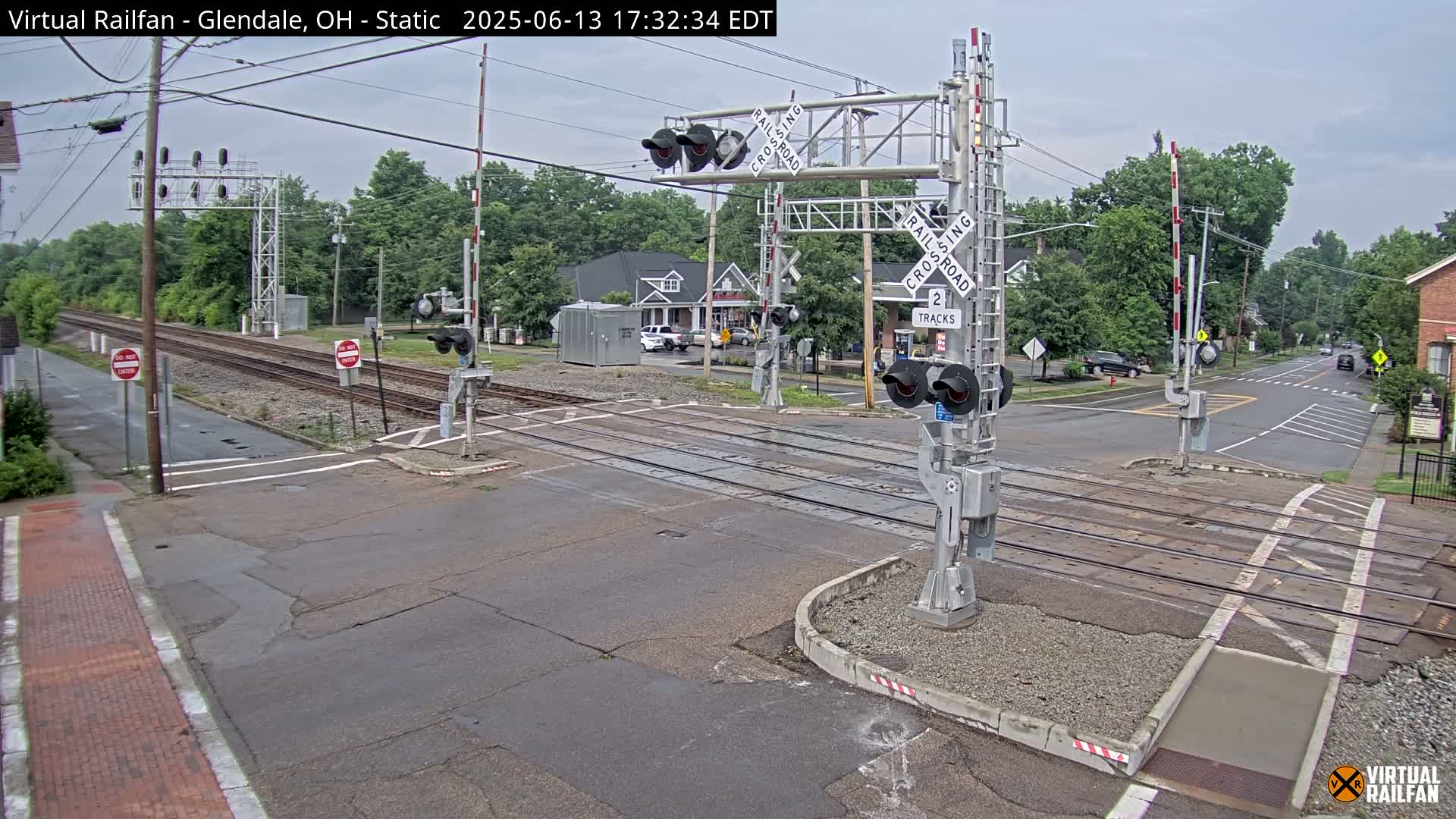 A railroad crossing with lowered gates is shown on an overcast day;  cars are visible on a nearby street.
