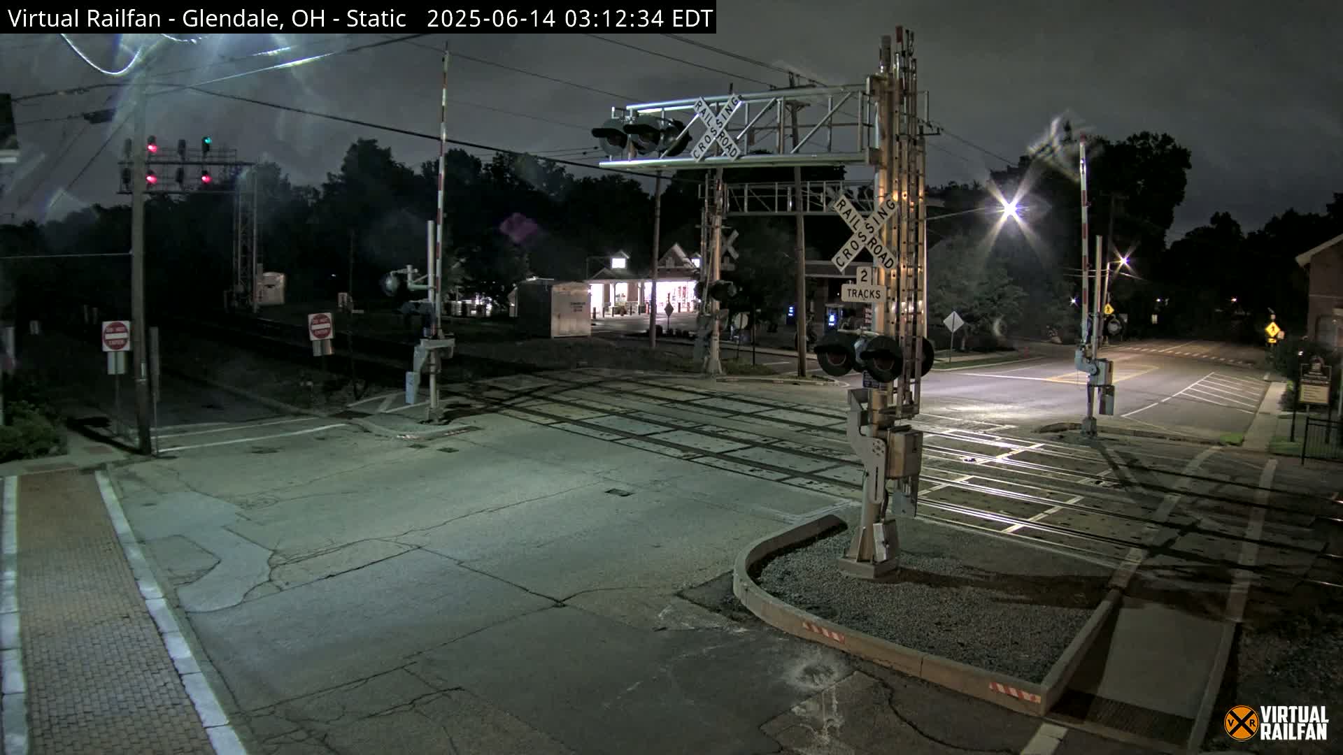 A nighttime view of a railroad crossing with its gates down, illuminated by streetlights and featuring a two-track crossing sign.