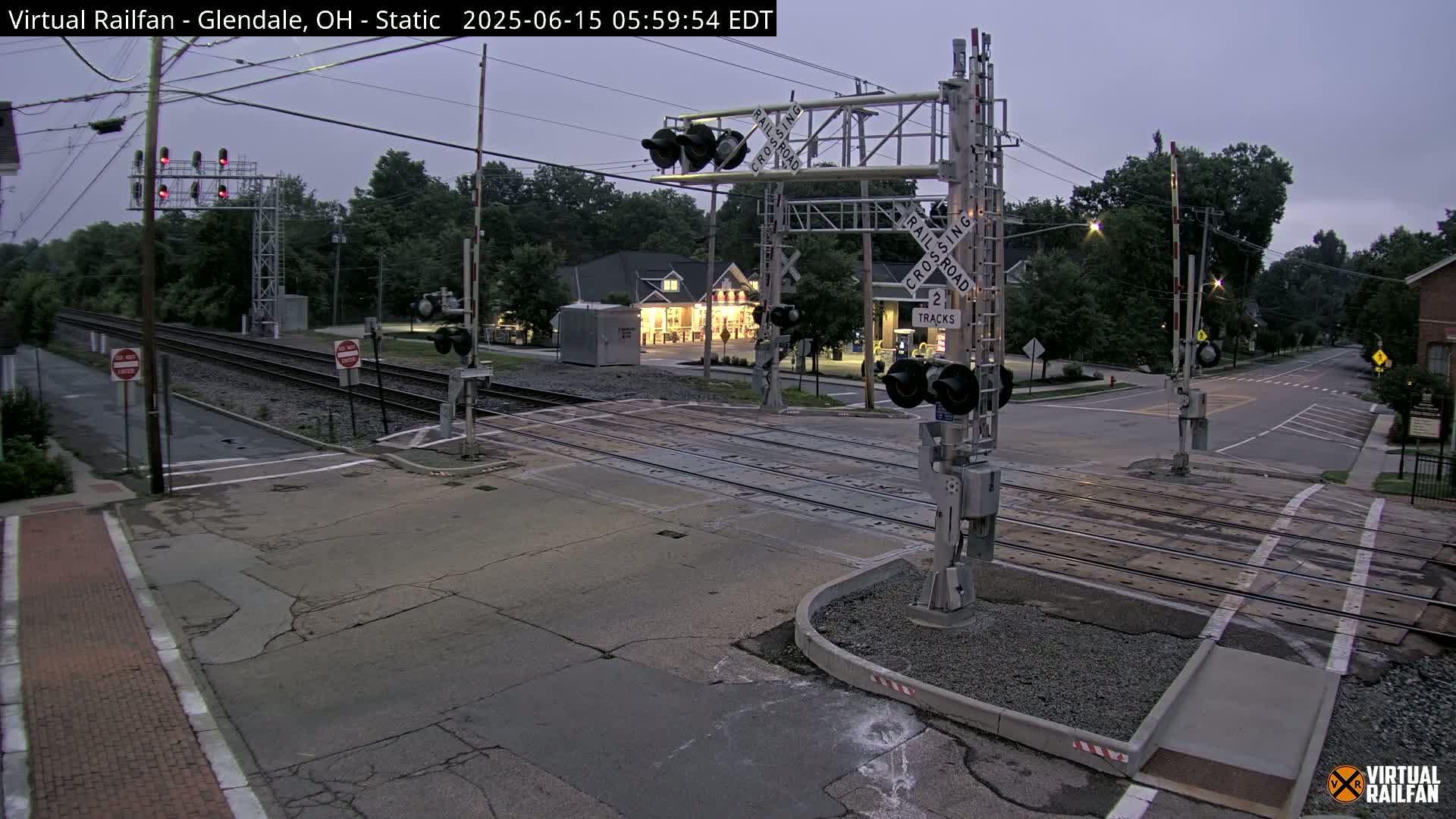 A railroad crossing with lowered gates under a dusky sky shows two tracks intersecting a road in a residential area.