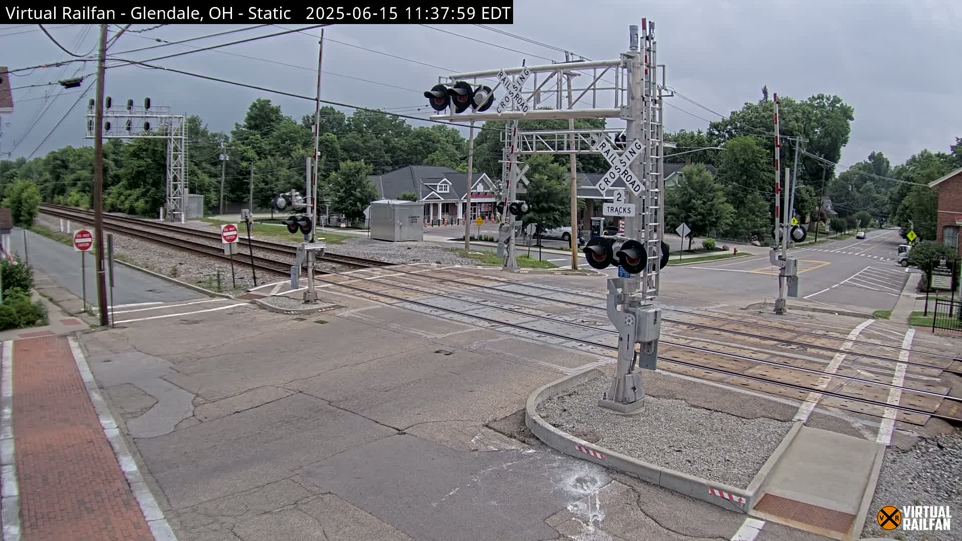 A railroad crossing with lowered gates is shown on an overcast day, with a road intersecting the tracks and buildings visible in the background.