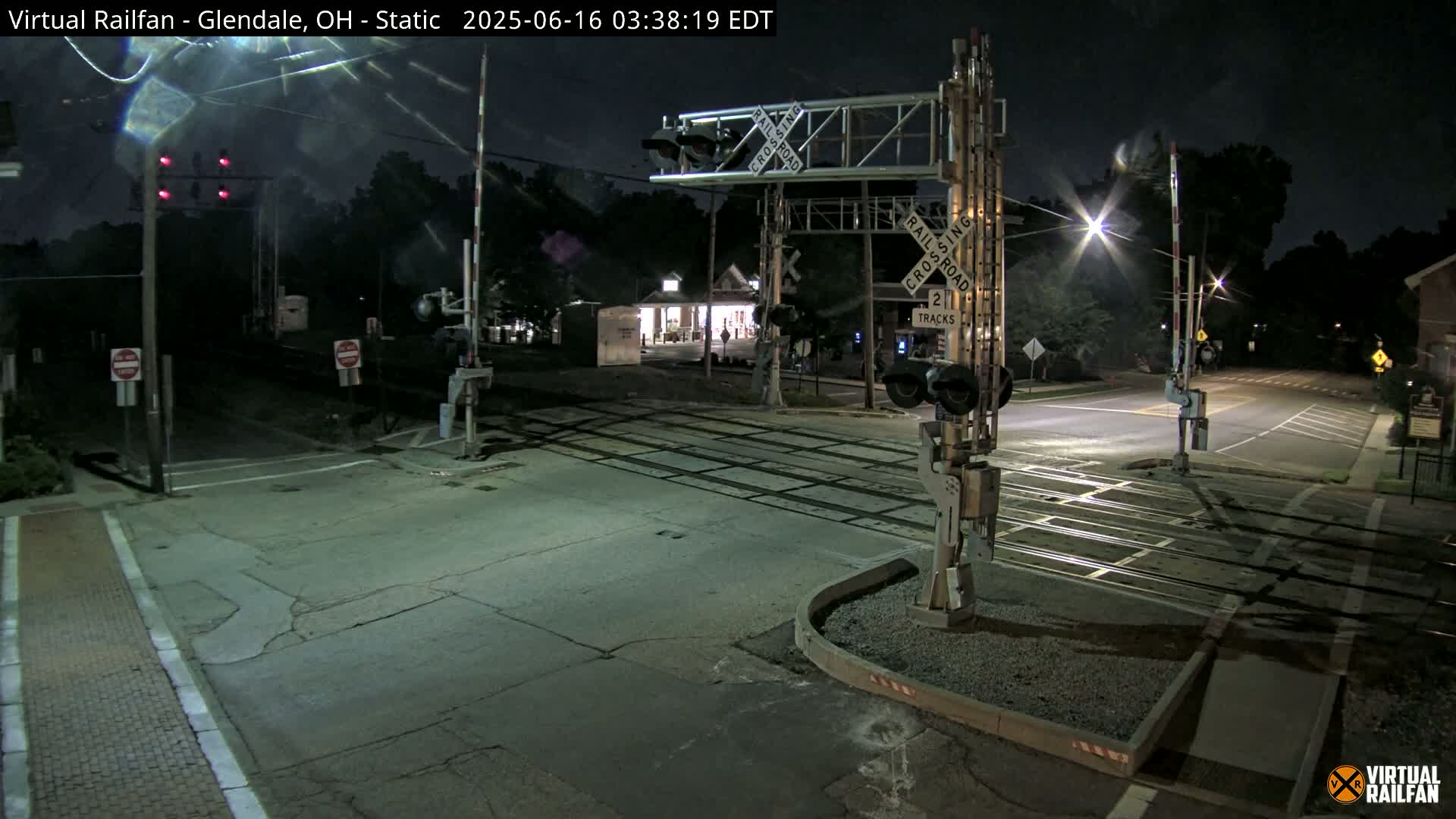 A nighttime view of a railroad crossing with lowered gates and red flashing lights, illuminated by streetlights.