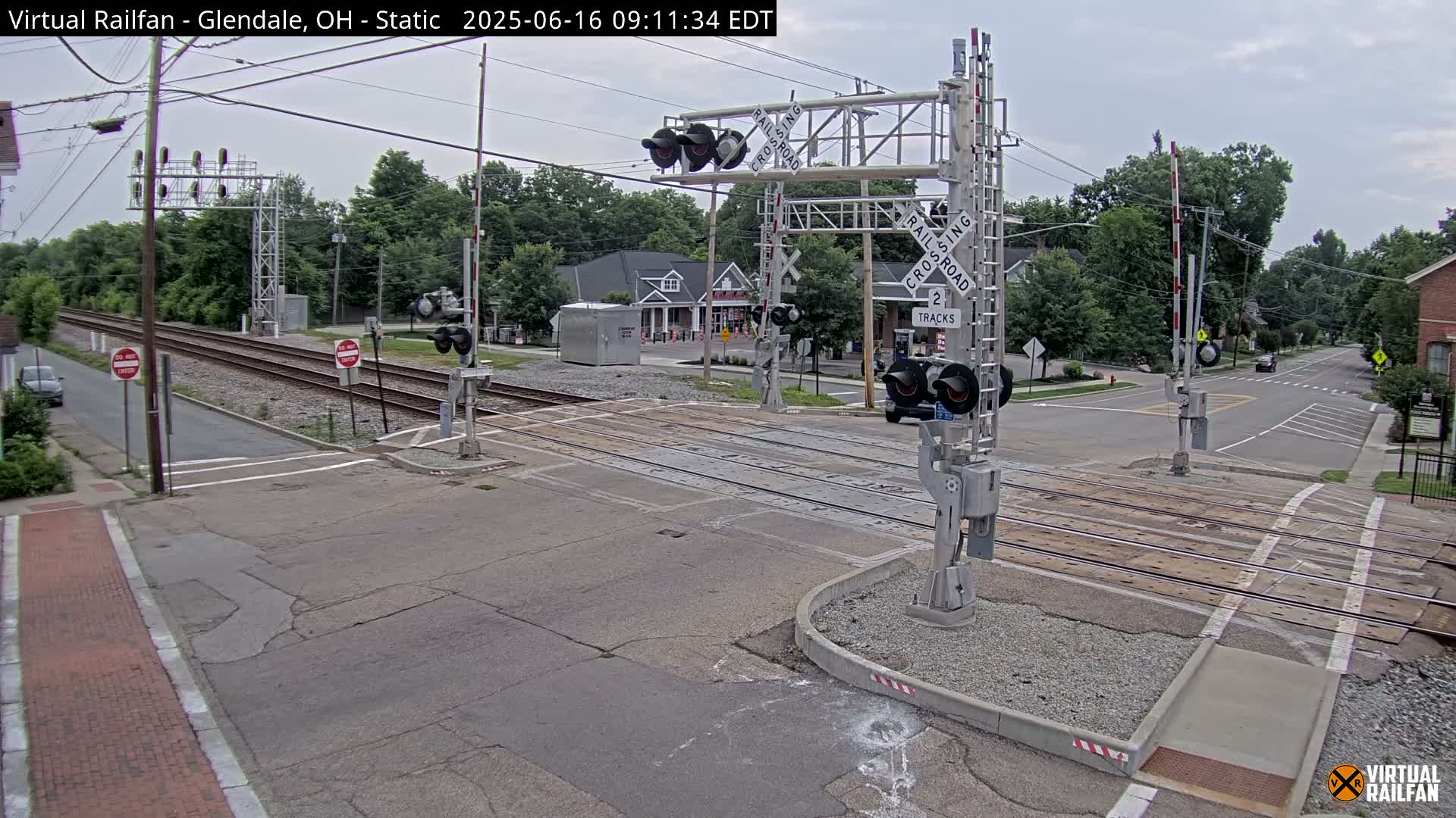 A railroad crossing with lowered gates shows two tracks intersecting a street under an overcast sky.